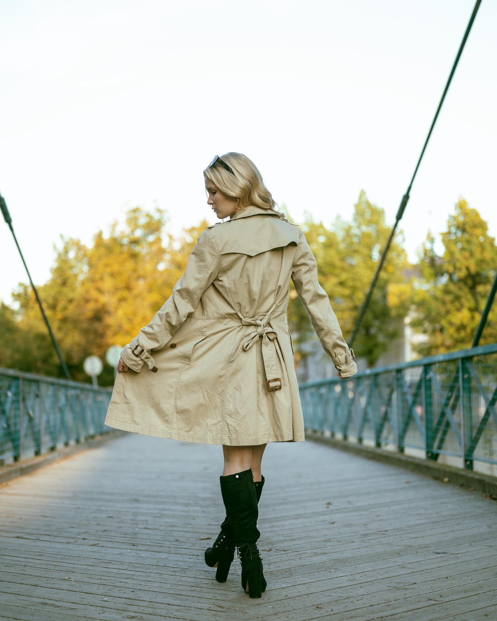 A person with long hair, wearing a beige trench coat and black knee-high boots, walking away on a wooden footbridge. The bridge has blue metal railings and is surrounded by trees with green and yellow foliage. The sky appears clear.