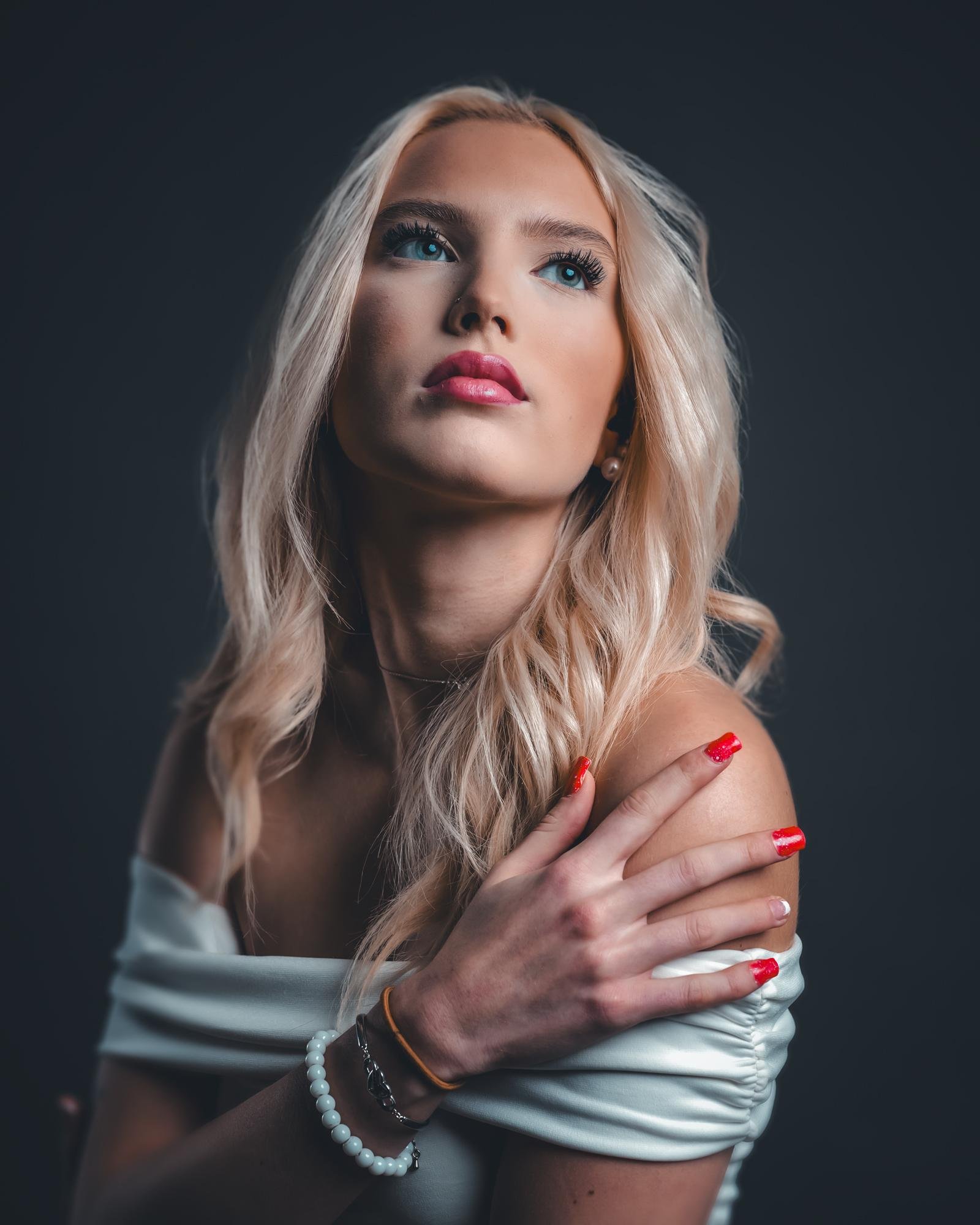 The image shows a person with long, blonde hair looking upwards. They are wearing an off-shoulder white top and have noticeable red nail polish. The lighting focuses on the individual's face and upper body, creating a dramatic effect with a dark background. The person is also wearing bracelets on one wrist.