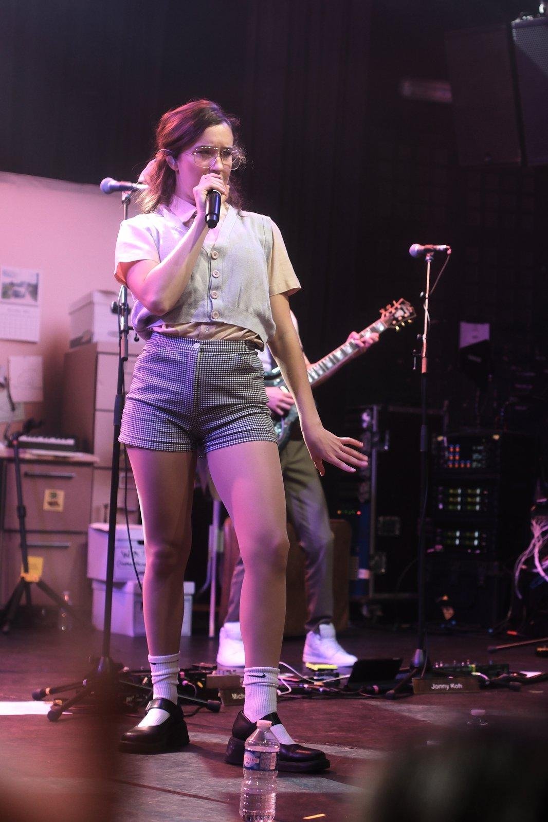 Photo verticale. Une jeune femme sur scène, habillée d’une chemise à manche
courte, un gilet gris sans manche, un short quadrillé noir et blanc, des
chaussettes blanches et des chaussures noires. Elle tiens un micro de la main
droite et chante dedans, la main gauche faisant un mouvement de danse. Elle est
éclairée par une lumière orange et rose

