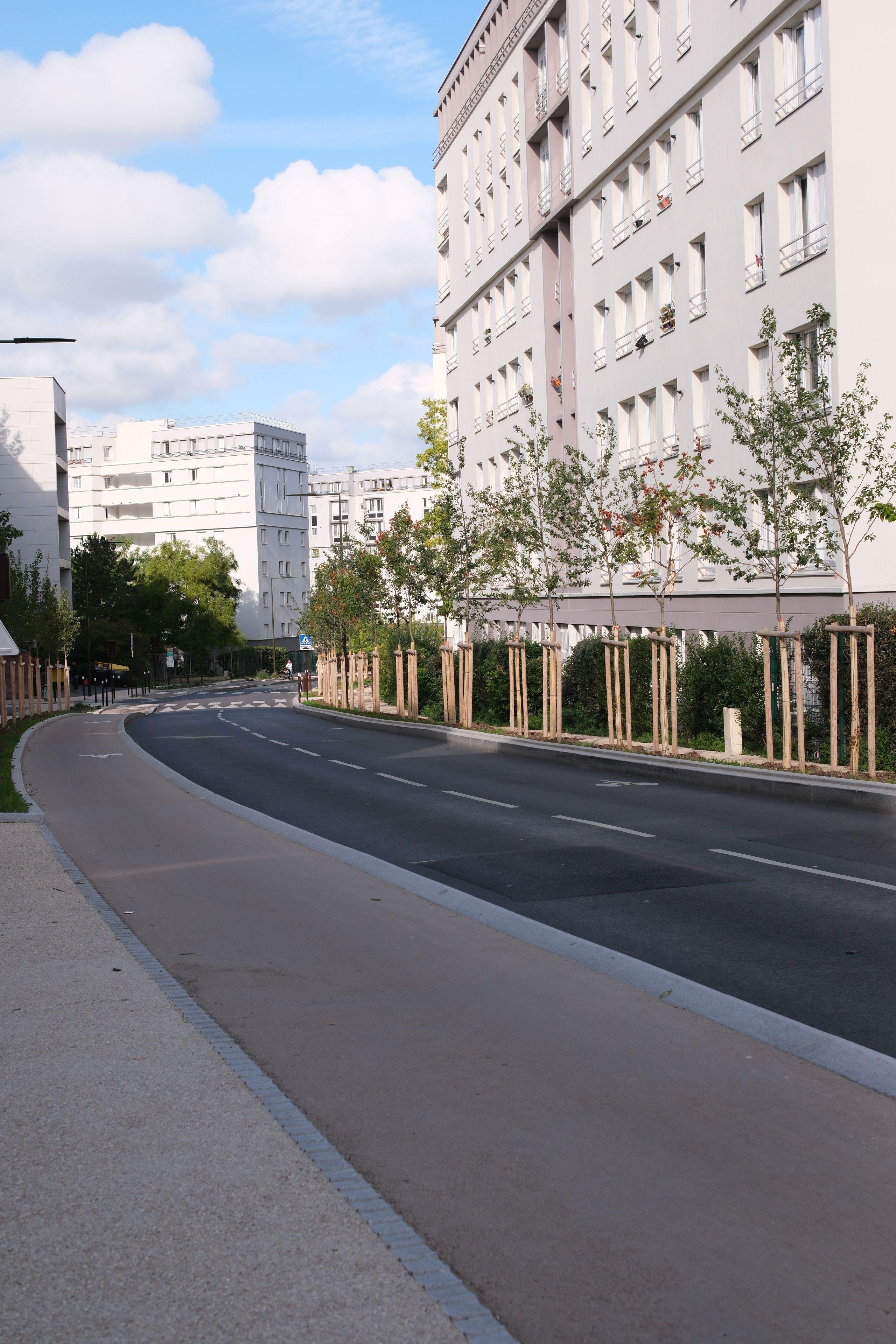 Photo verticale. Une Route à 2 voies et qui va légèrement sur la droite, entre 2 pâtés d’immeubles modernes. Des jeunes arbres en bordure, une piste cyclable au premier plan et qui suis la route, sous un ciel bleu avec des nuages
