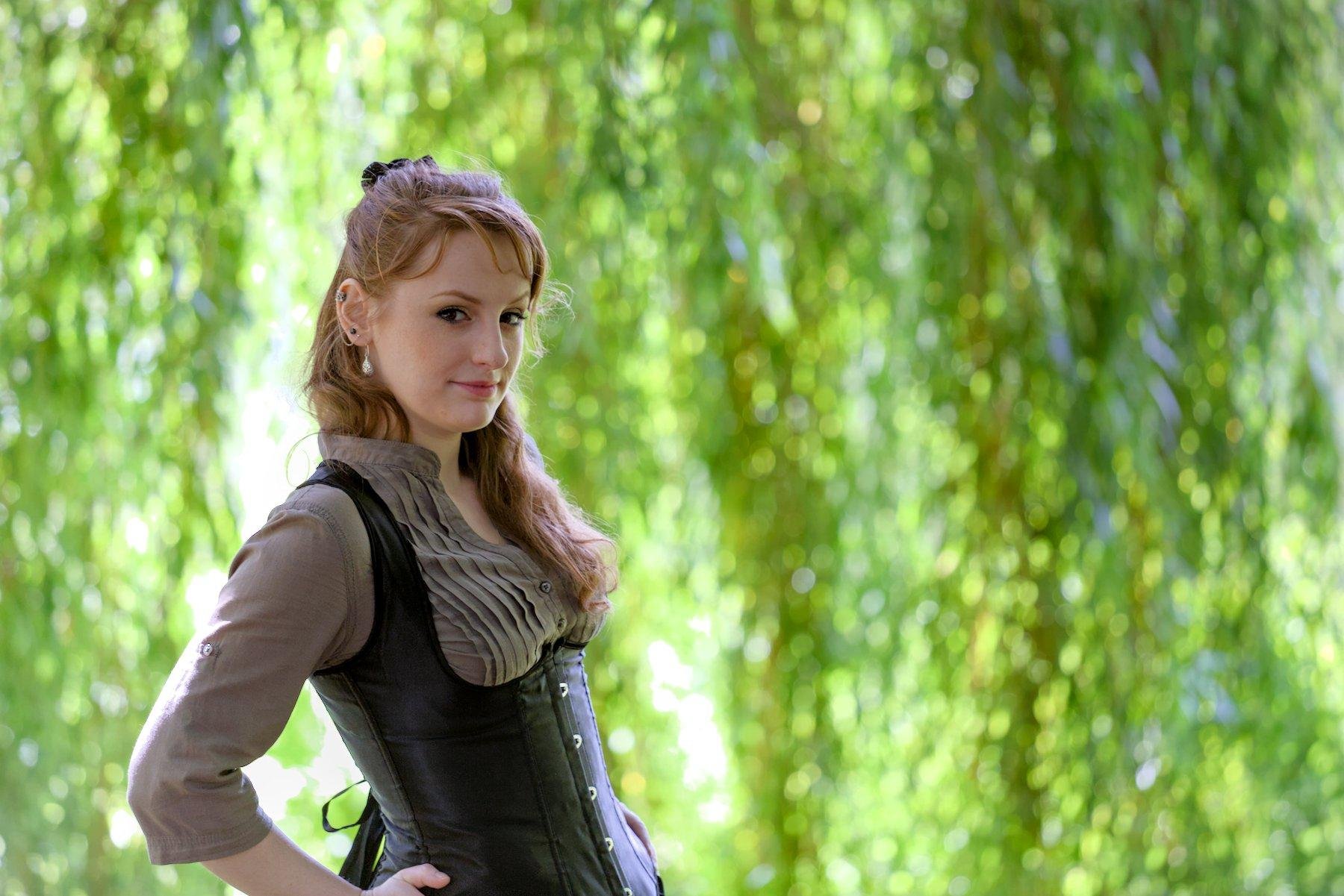 Photo horizontale. Une jeune femme rousse au cheveux long élégamment coiffé, portant un chemisier marron et par dessus, un corset noir. Elle se tiens debout, sa main gauche sur la hanche, légèrement penchée en arrière. Le fond est composé de feuillages verts