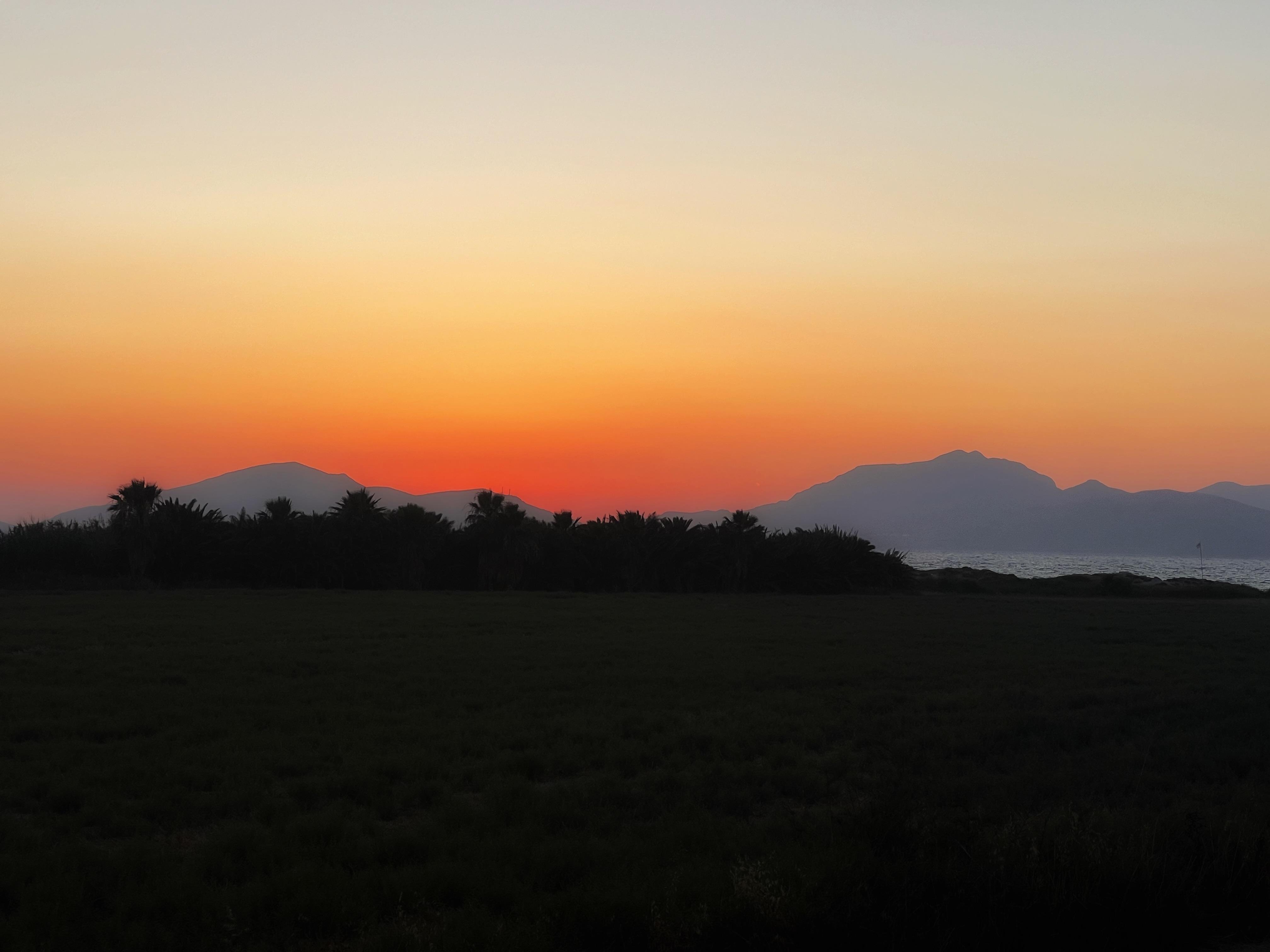 Dark grassy field in the foreground with a line of silhouetted trees, distant mountain shapes on the horizon, and a calm sea at right beneath an orange-to-white sunset sky.