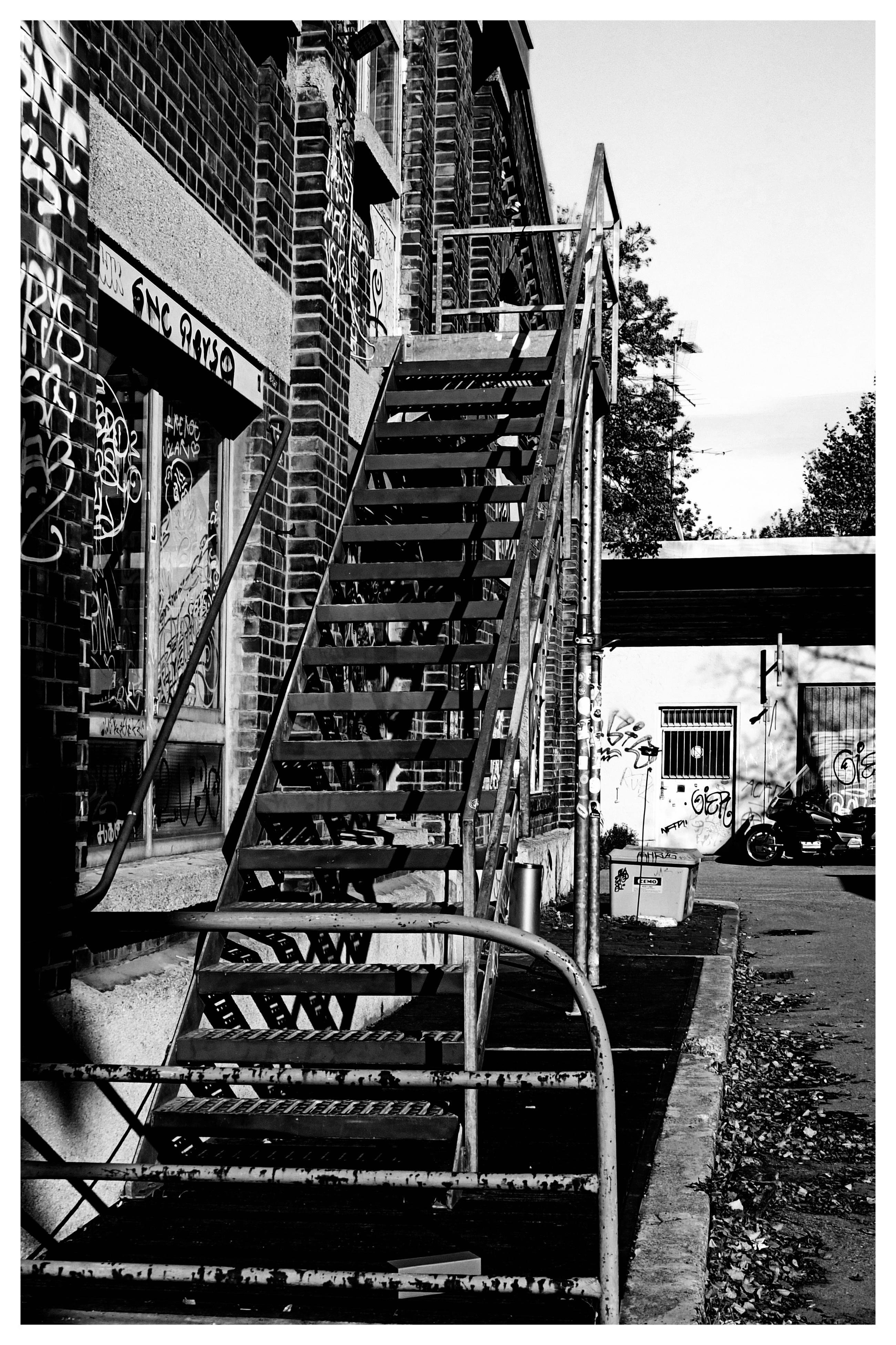 Black-and-white view of a metal exterior staircase attached to a brick building covered in graffiti, with steps and railings rising diagonally from the foreground to an upper landing; an alley and a smaller graffiti-marked building appear in the background.