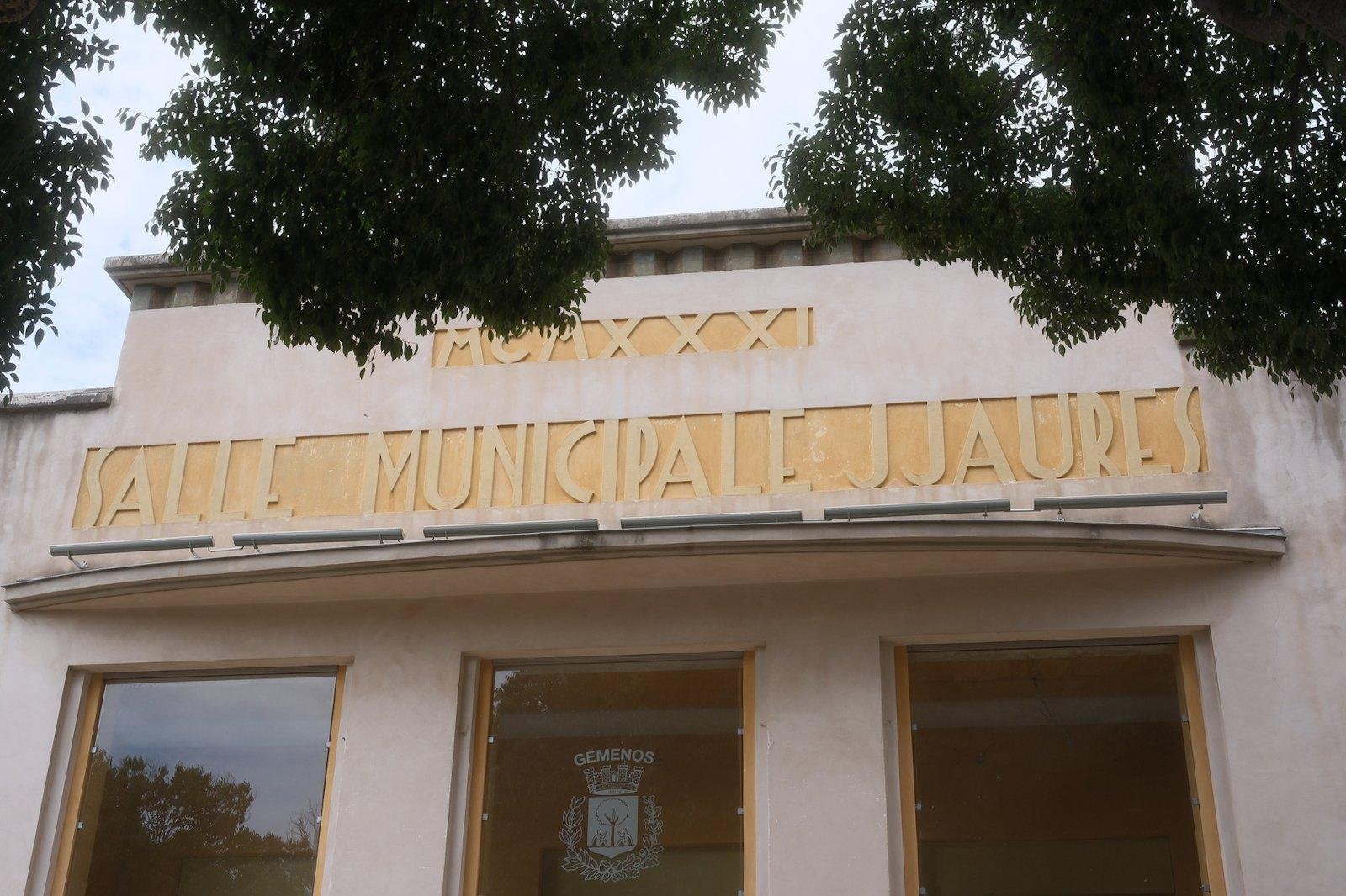 Horizontal photo. A building with the top having markings saying “MCMXXXI SALLE MUNICIPALE JJAURES” in a Art Nouveau style of font. Below a sign is engraved in a window saying “Gemenos” and the city’s symbol a stylised castle and below a tree in a shield surrounded by some plant. The frame is invaded by gree tree leaves at the top