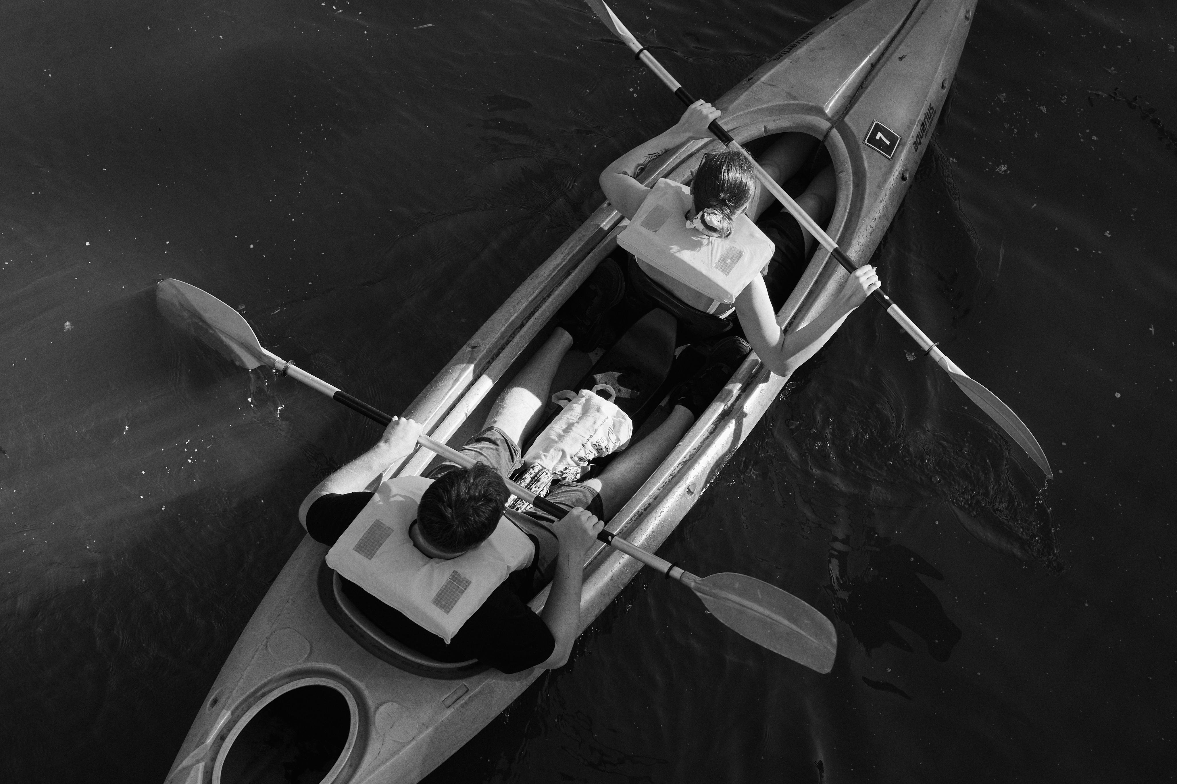 The image is a black-and-white photograph of two people in a tandem kayak. They are paddling on a body of water. Both individuals are wearing life jackets and holding paddles, with one person seated in front and the other in the back. The photo is taken from an overhead perspective. The water surrounding the kayak appears calm.
