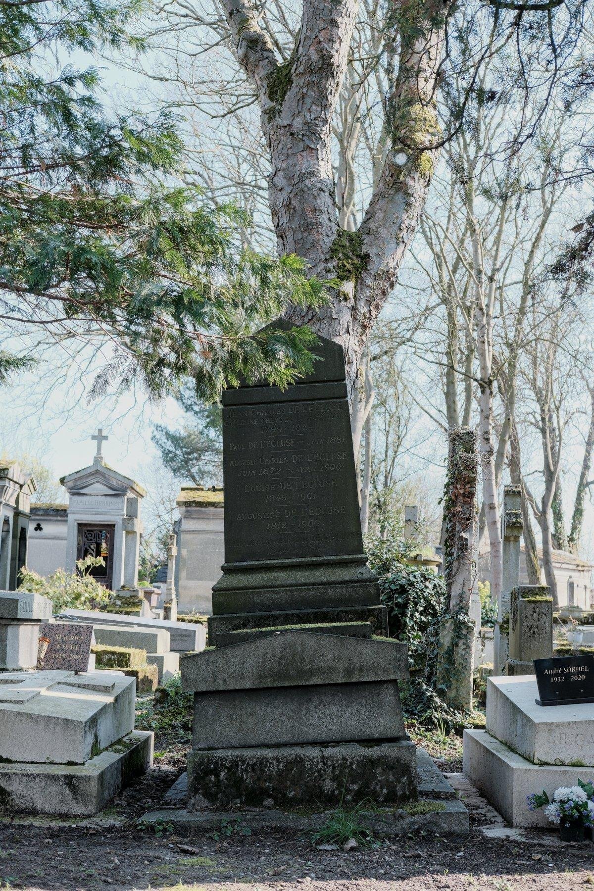 Photo verticale. Une tombe avec une grande stèle au dessus. La pierre tombale est légèrement penchée vers la gauche, comme si on avait tenté de l’ouvrir…