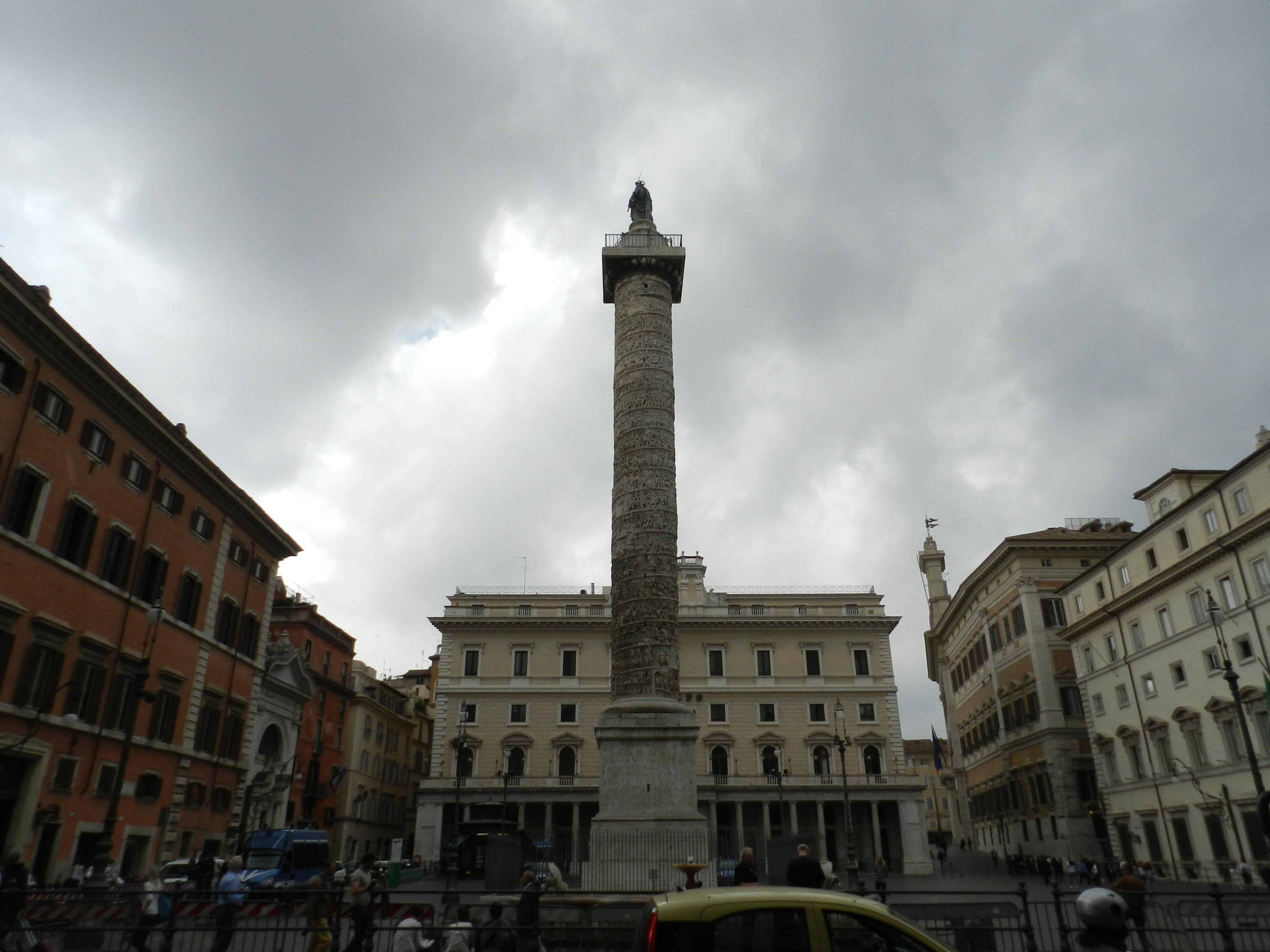 A wide view of the Column of Marcus Aurelius rises at the center of Piazza Colonna in Rome, Italy, framed by historic buildings under a dramatic, overcast sky. The tall marble column, carved with an intricate spiral frieze depicting Marcus Aurelius’s campaigns against the Germanic tribes, stands on a sturdy square base. At its top is a bronze statue of Saint Paul, added in the 16th century to replace the original statue of the emperor. In the background stands the elegant Palazzo Wedekind, with its neoclassical façade of arched windows, columns, and a central clock. Surrounding the square are warm-toned Roman buildings, including ochre and cream façades with shuttered windows. At street level, pedestrians, cars, and a scooter add a sense of busy Roman city life, contrasting with the ancient monument’s enduring presence.
