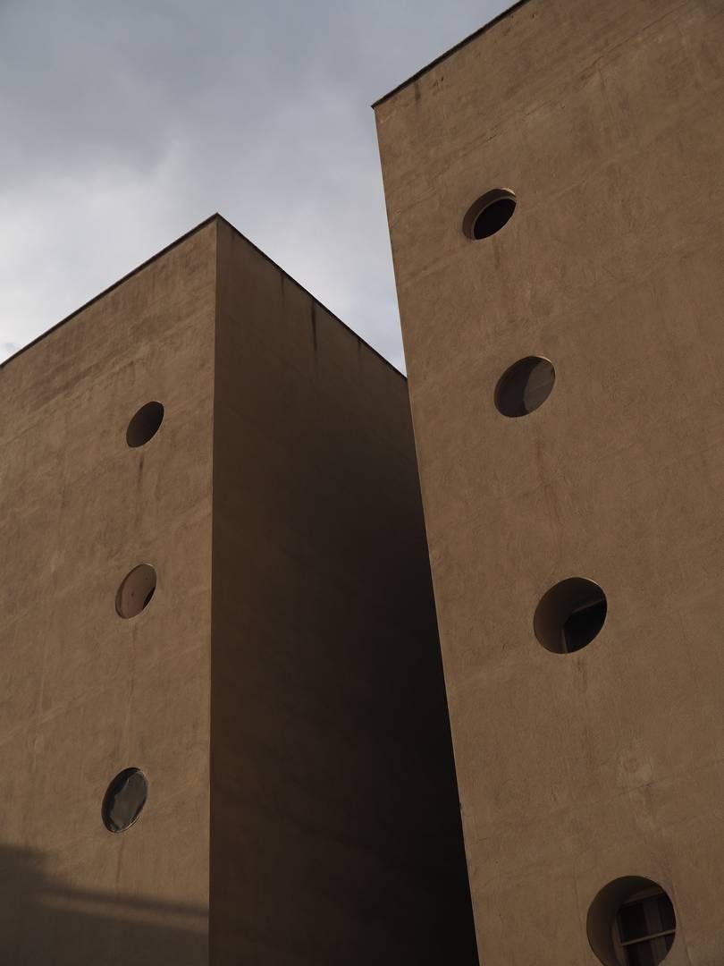 Due palazzi affiancati, separati da uno spazio di pochi metri. Le pareti sono forate solamente da piccole feritoie circolari, rinchiuse internamente da finestre. Il cielo è nuvoloso.