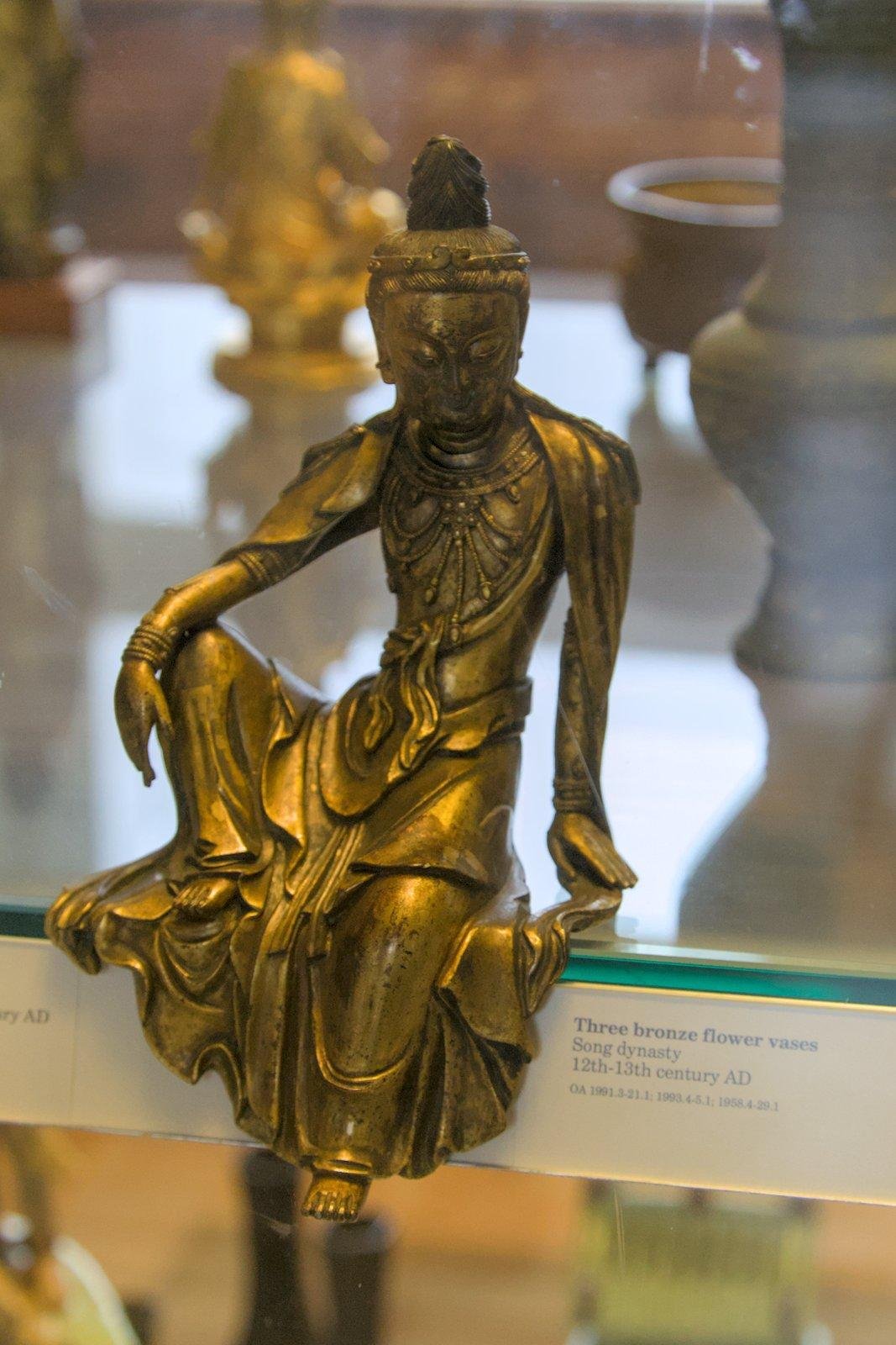 Photo format portrait montrant une statuette dorée d’un bouddha thaïlandais je crois, assis sur le bord d’un plateau de verre d’une vitrine de musée