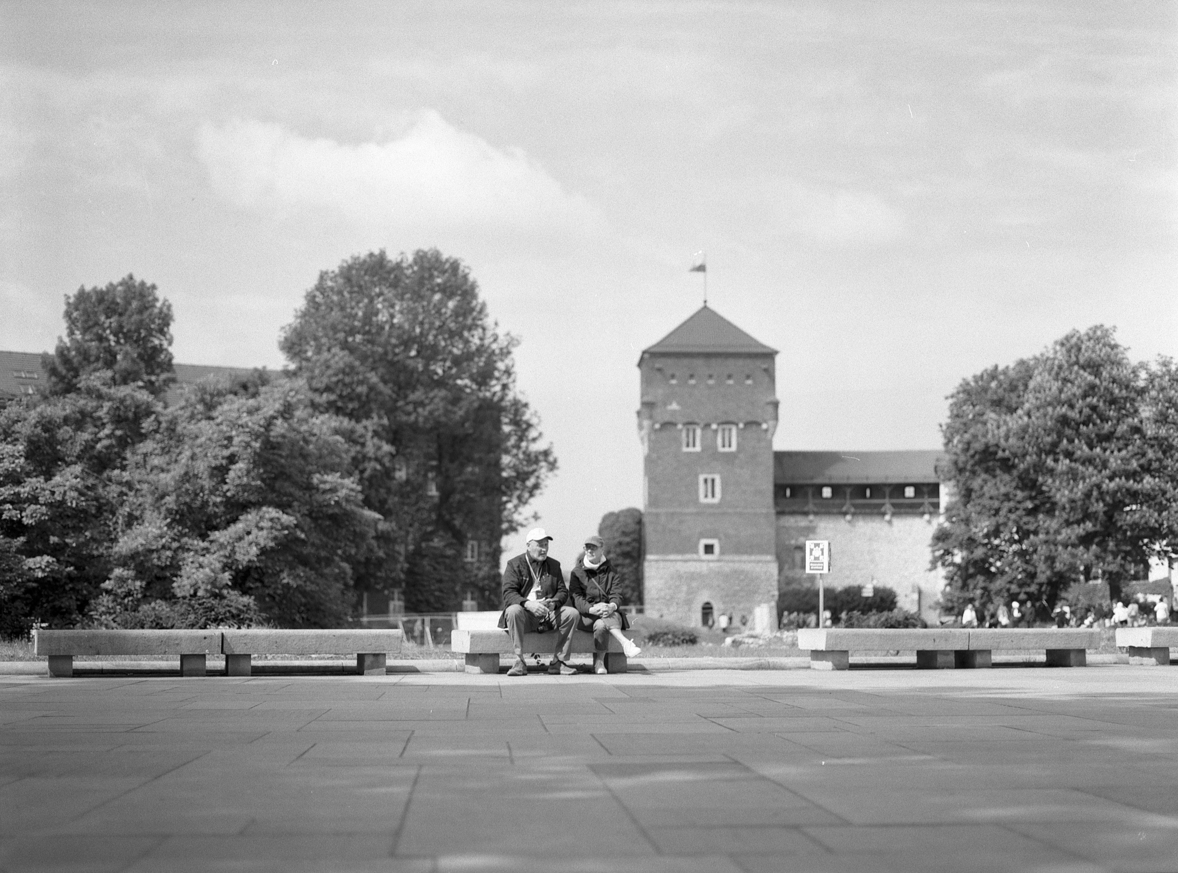 Two people sitting on a bench in a spacious, paved plaza with large leafy trees and a prominent brick building featuring a tower and flag in the background. The scene is captured in black and white.