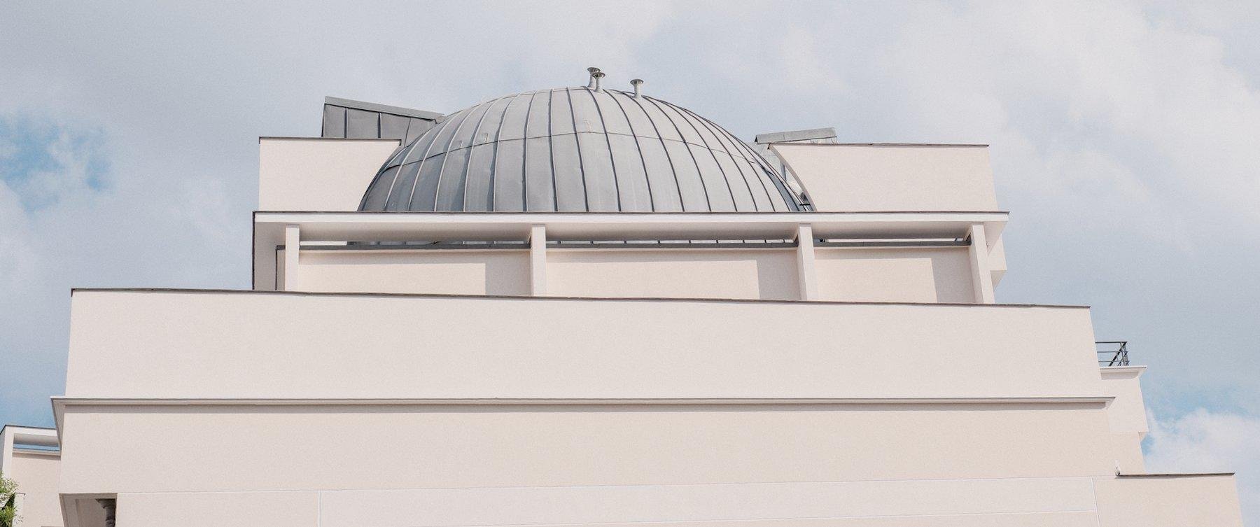 Photo horizontale. Au sommet d’un immeuble beige en étages de plus en plus petit, formant un podium, un dome gris