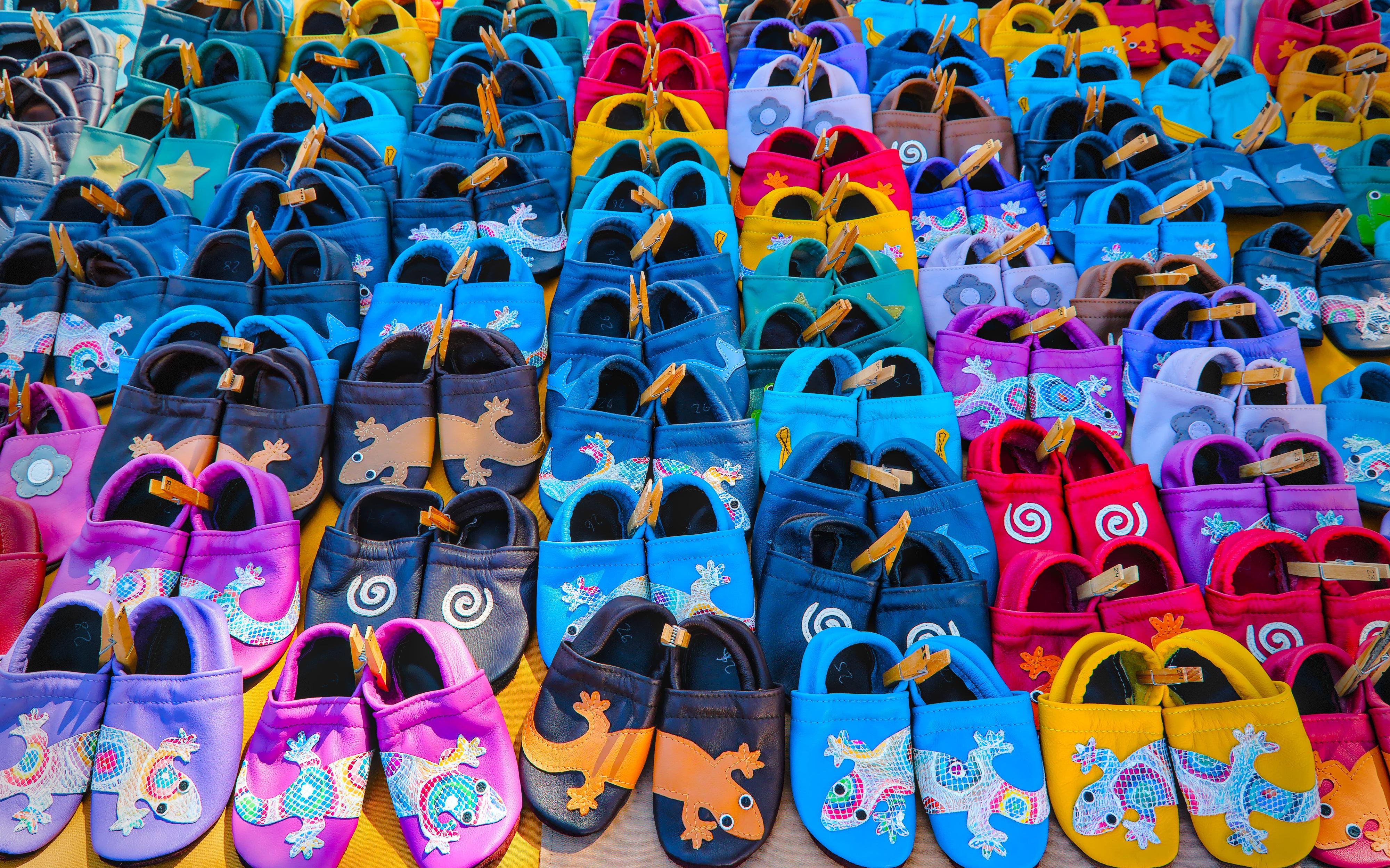 Rows of colorful leather slip-on shoes arranged tightly together, featuring embroidered designs like geckos, flowers, stars, and spiral patterns in blue, pink, yellow, and purple.
###
Reihen aus bunten Leder-Slip-on-Schuhen, die eng miteinander verbunden sind, mit bestickten Designs wie Geckos, Blumen, Sternen und Spiralmustern in Blau, Rosa, Gelb und Lila.