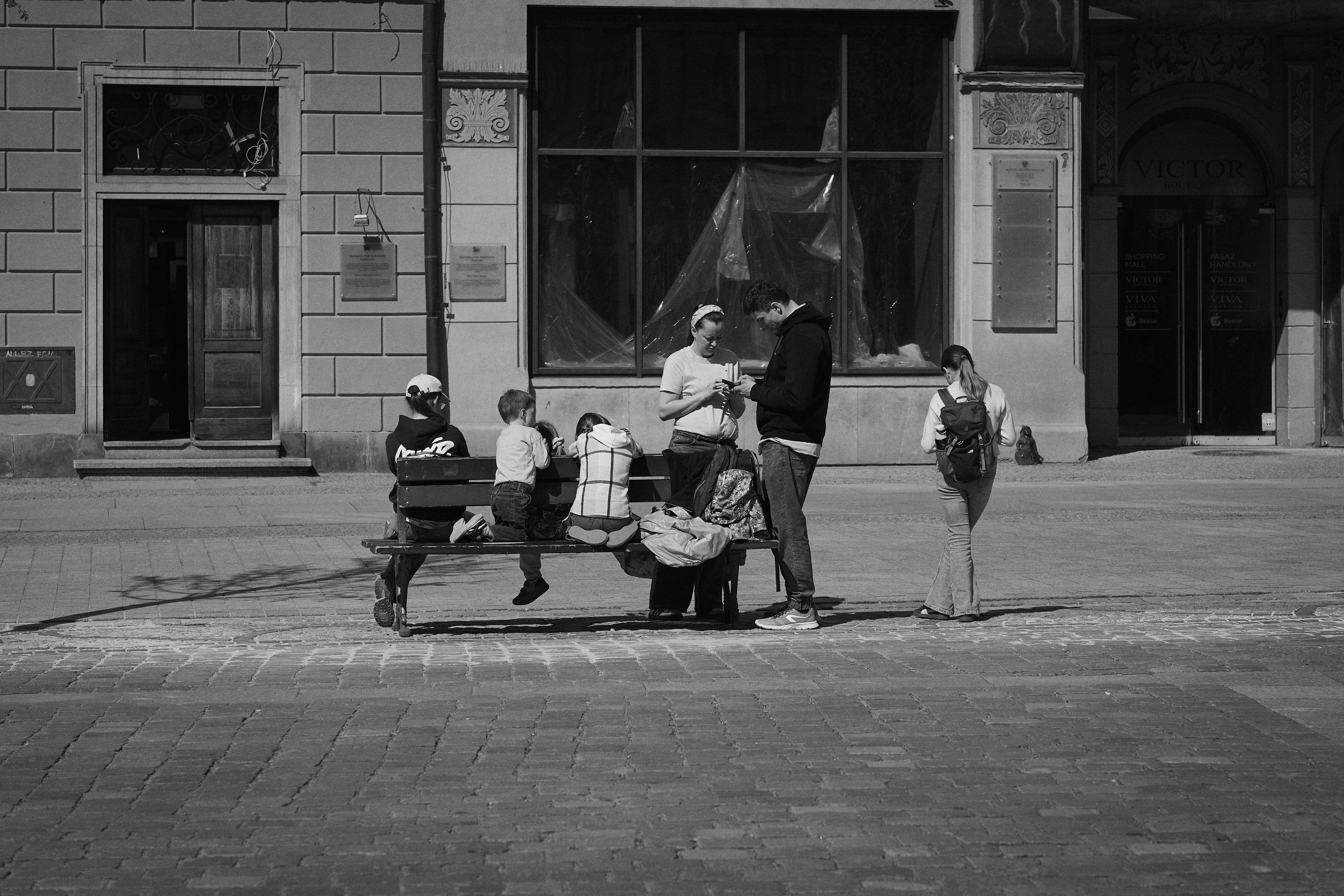 A black-and-white street view with a bench in a cobblestone square. Three children sit on the bench, two adults stand in front, staring at their phones. Another person stands to the right, near a shop door and large windows.