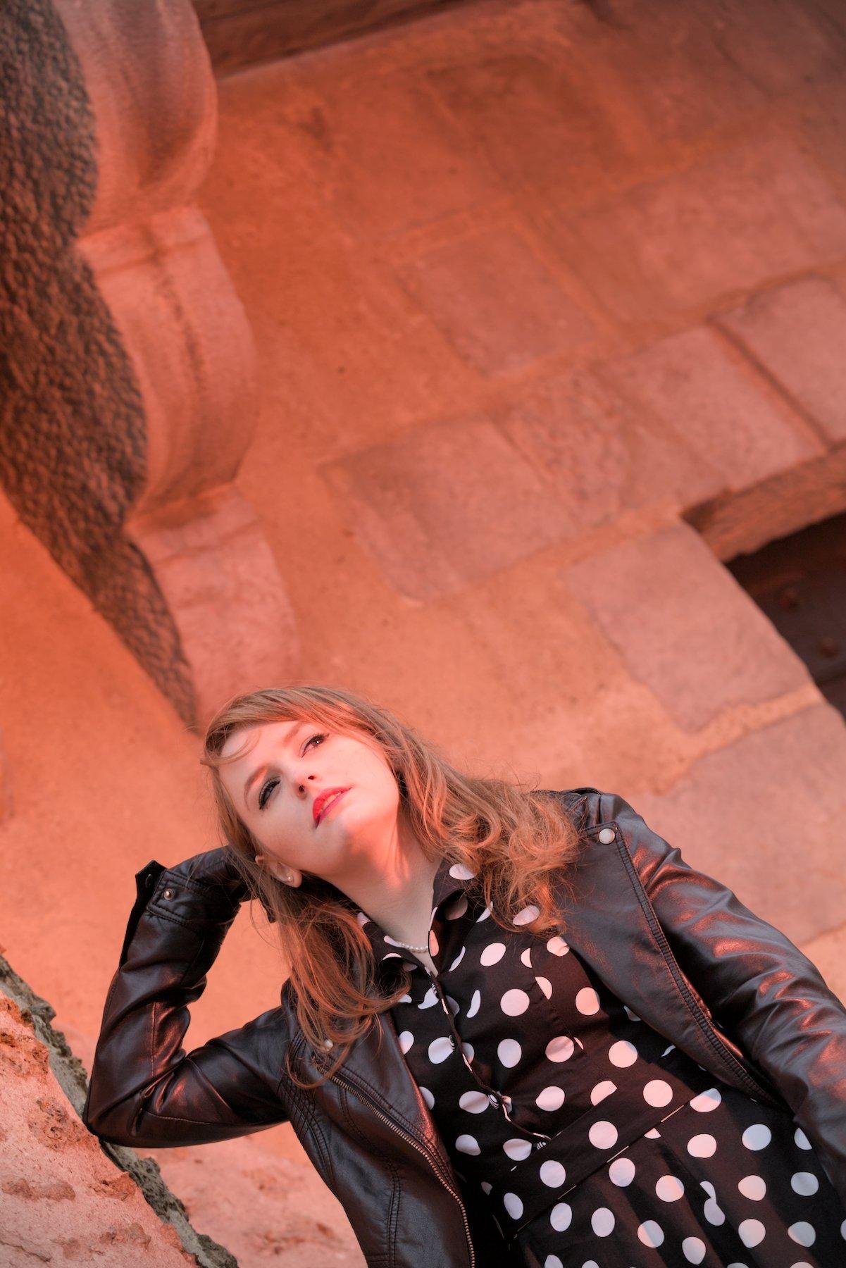 Photo verticale, penchée. une jeune femme rousse une robe noire à pois blanc genre années 50, une veste en cuir noir et des gants noirs en cuir. Elle a son coude droit appuyé contre un vieux mur en pierre et sa tête posée sur le dos de sa main droite, regardant droit devant elle