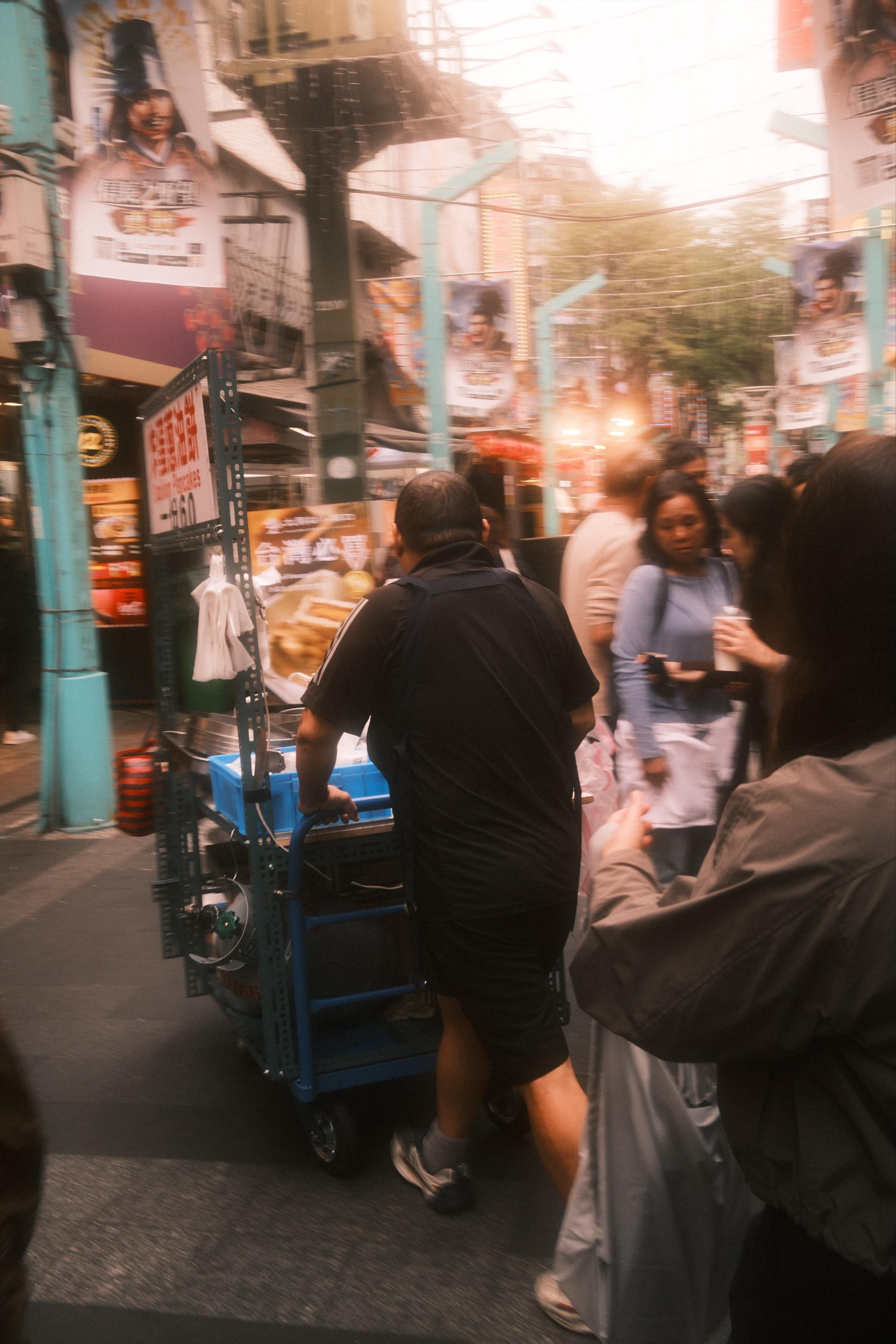 Crowded city street with a man pushing a blue handcart through pedestrians; storefront signs and posters line both sides, with overhead wires and a bright, hazy light in the distance.