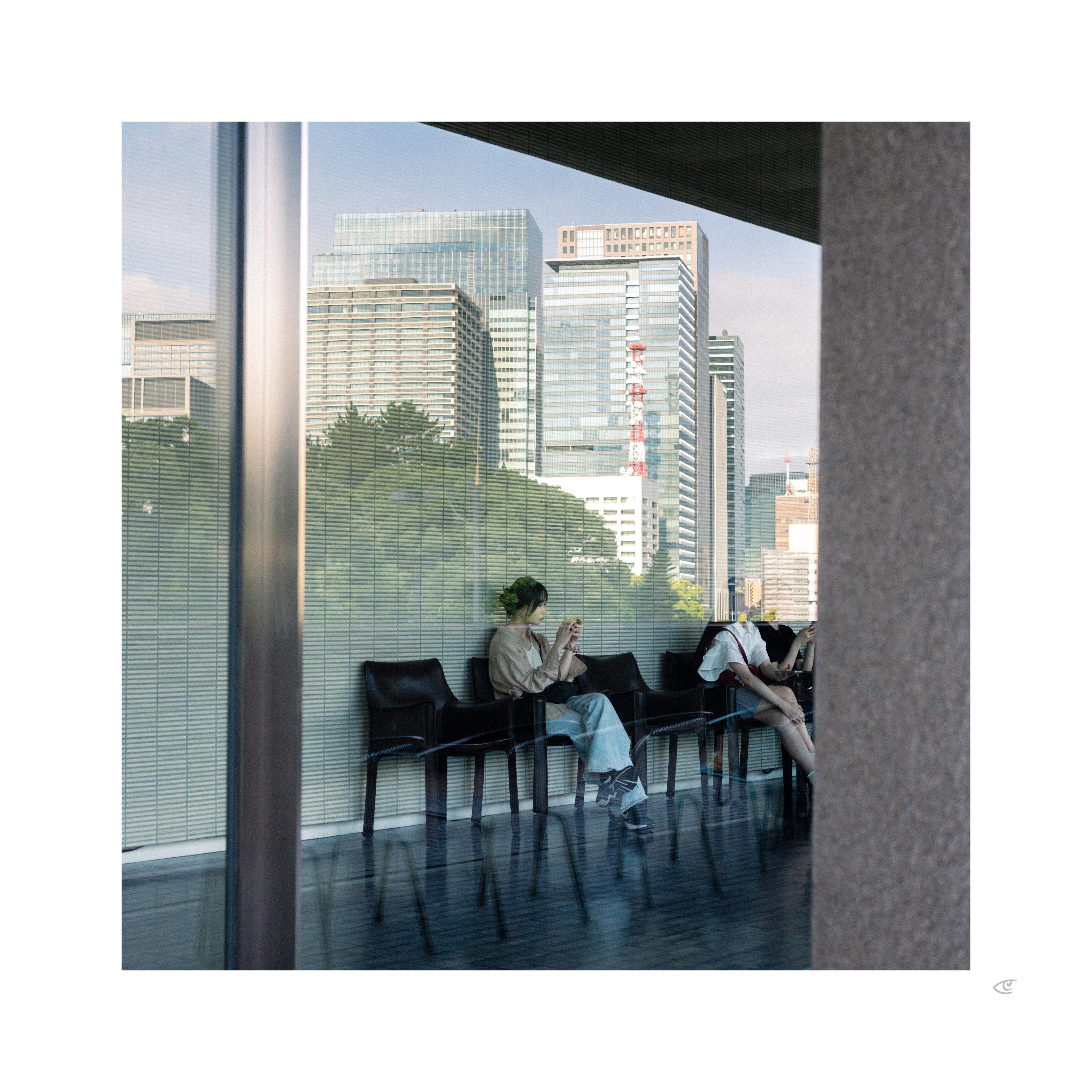 Two people seated on black chairs in a glass-walled lobby, viewed through a reflective window; one looks at a phone while the other leans forward. City skyline with tall office buildings and a red-and-white tower visible outside, with a vertical metal frame and a concrete column bordering the right edge.