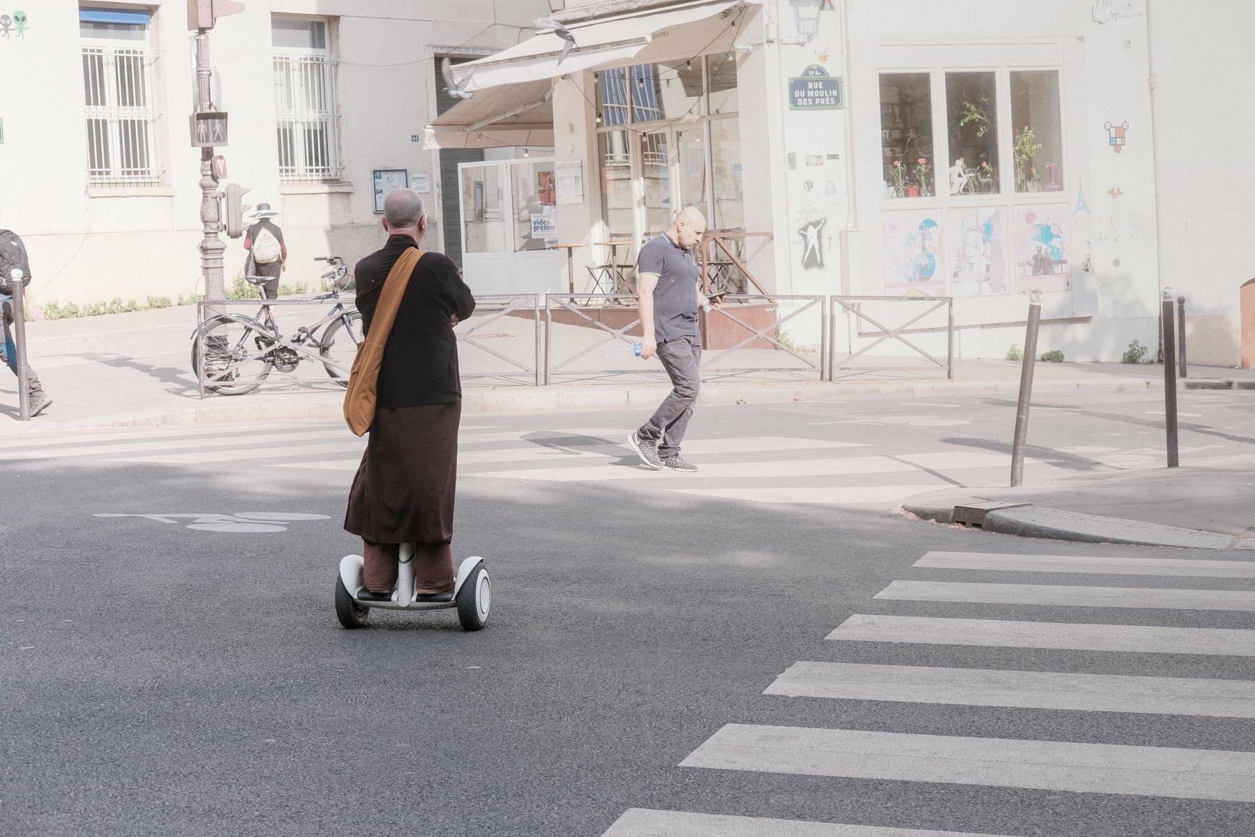 Photo horizontale. Un homme de dos, en pantalon marron, longue toge marron, manteau marron, un sac en bandoulière marron clair, aux airs de bonze, sur un genre de hoverboard, traverse la rue, les bras croisés
