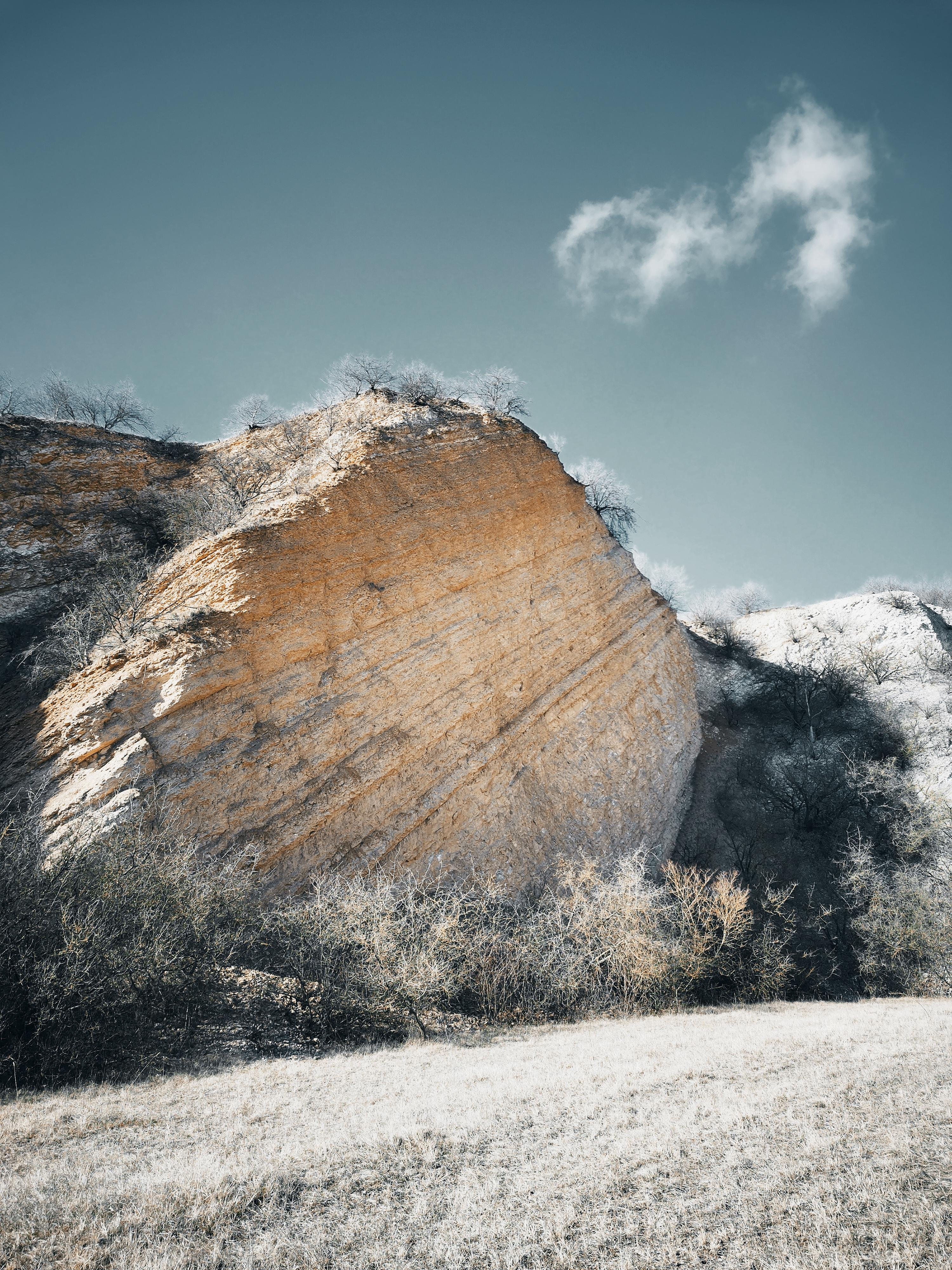 The image shows a large rock formation with an angled, rugged surface. It is surrounded by sparse vegetation, including bushes and small trees. The sky in the background is clear with a single fluffy cloud, giving the scene a peaceful and natural appearance.