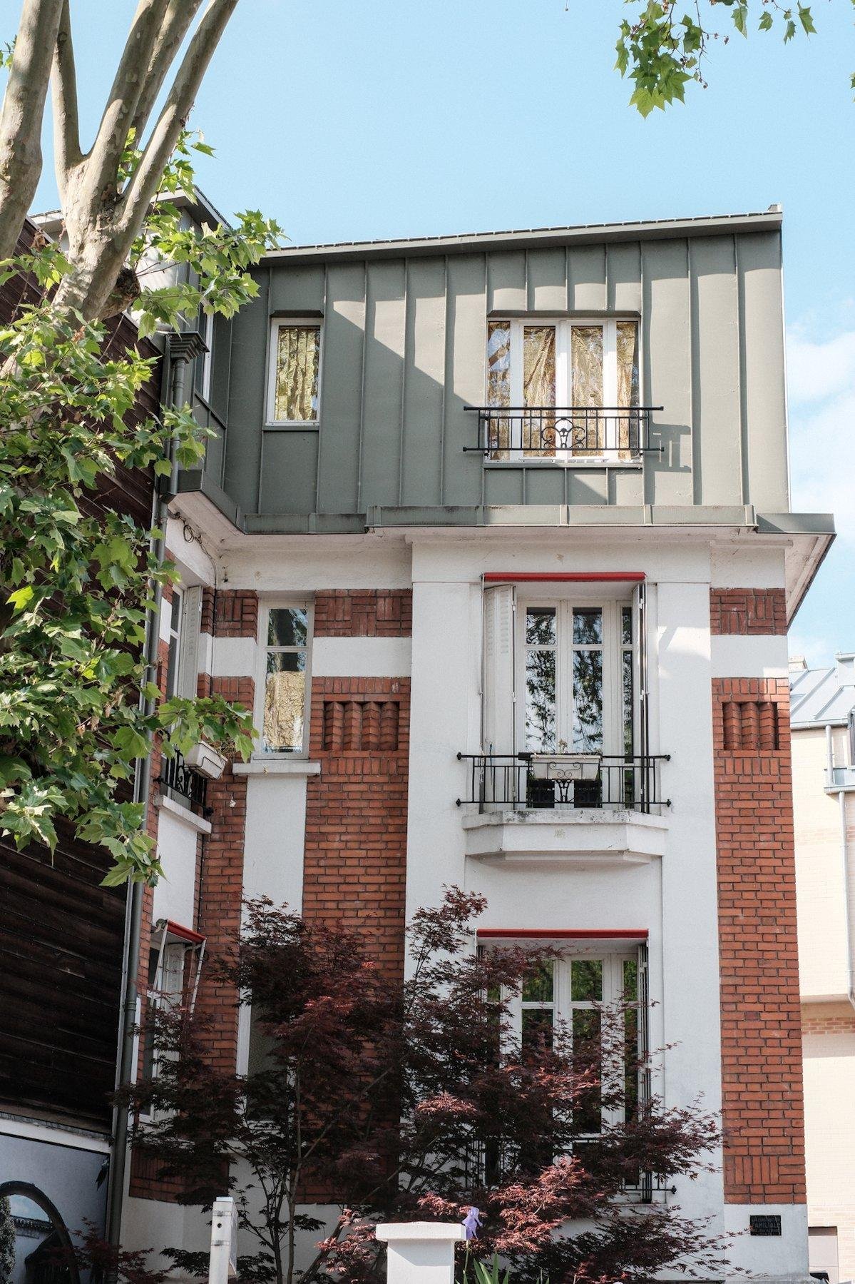 Photo verticale. Un immeuble en partie en brique rouge et béton peint en blanc, avec son dernier étage en tôle peinte en vert pâle