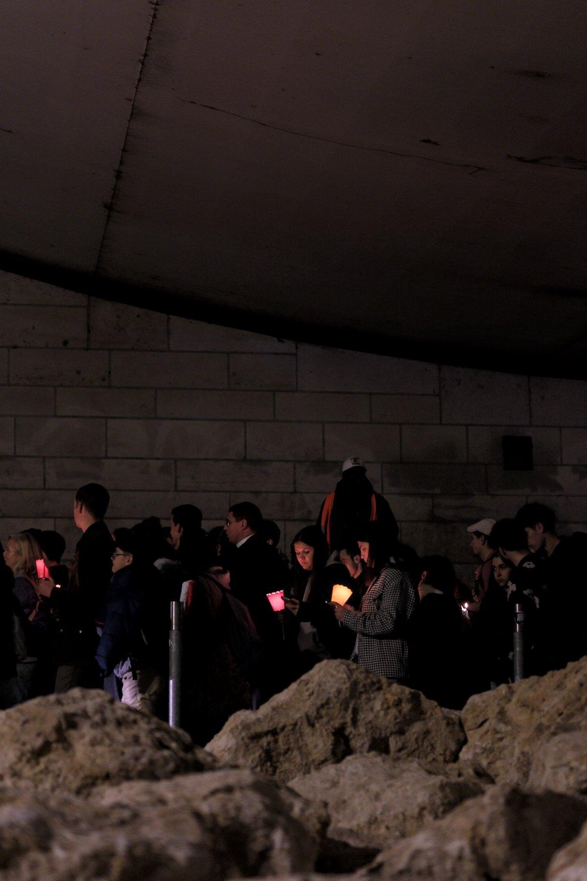 Photo verticale de nuit. Sous un pont, des gens avec tous une bougie allumée, vraie ou fausse, avancent vers la gauche de la photo