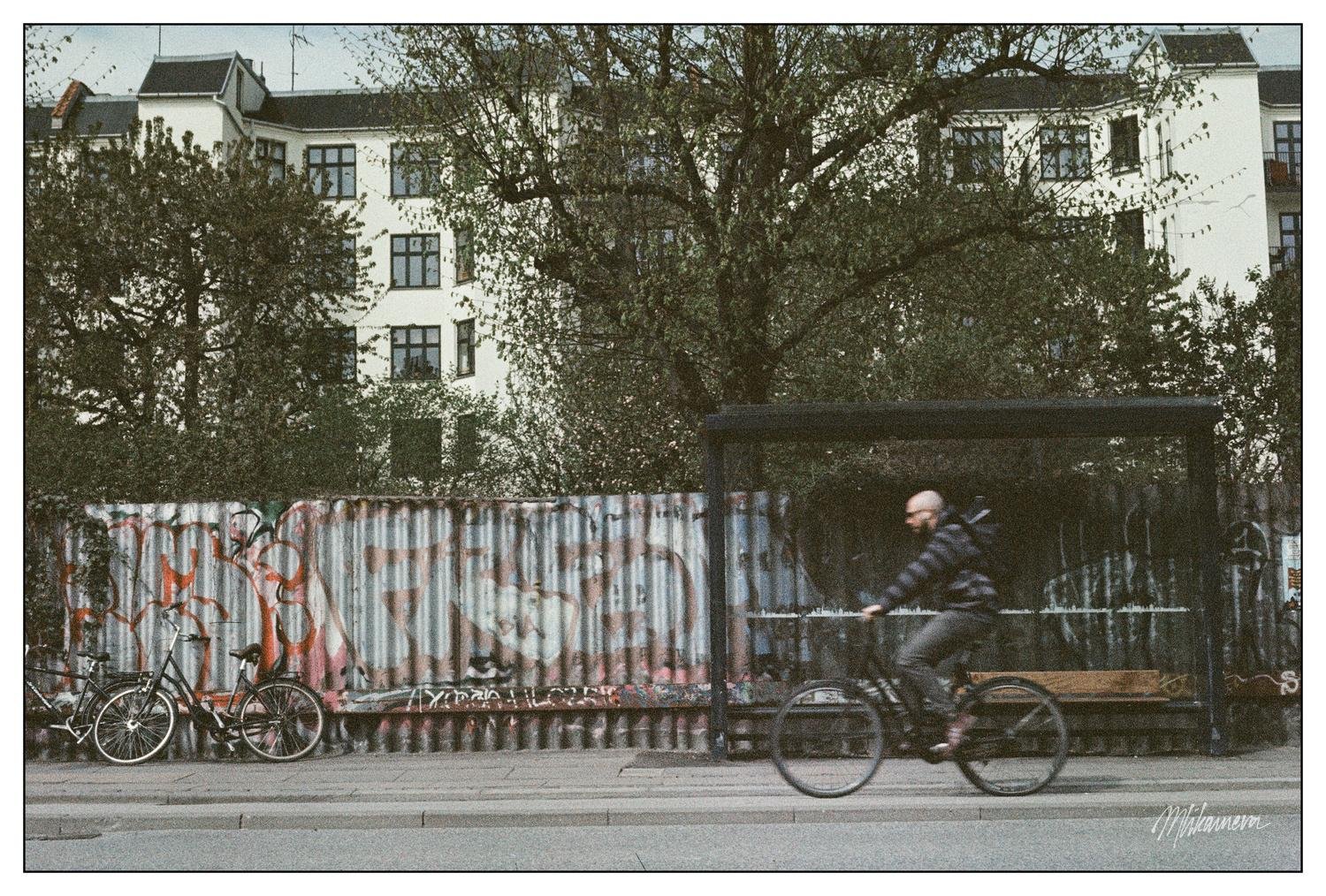 Cyclist riding past a graffiti-covered metal fence and a bus stop with a bench, surrounded by trees and multi-story buildings in the background. A few bicycles are parked by the fence, and the scene showcases an urban setting with a blend of greenery and architecture.