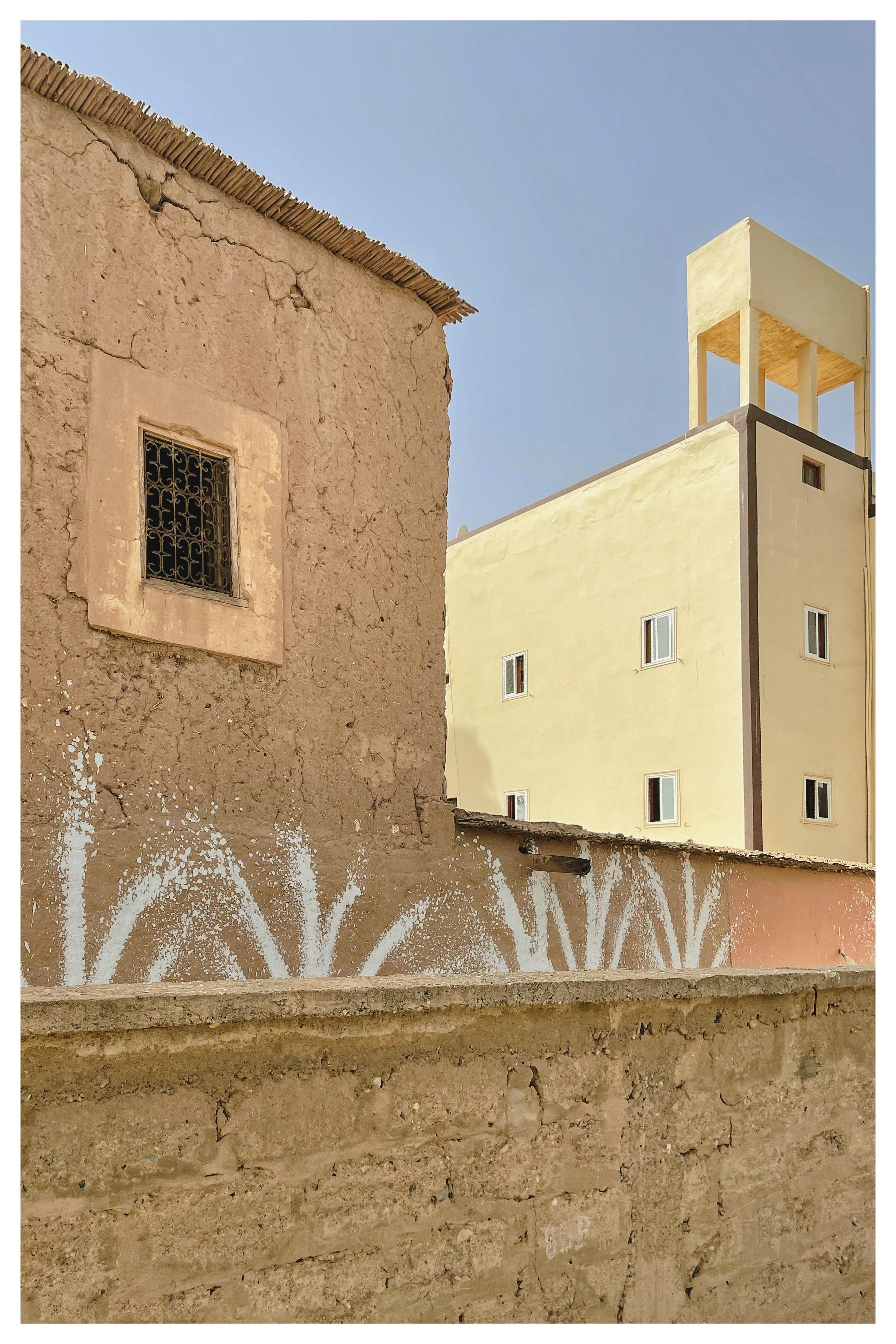 Adobe building with a small barred window, featuring cracked walls and a thatched roof edge. In the background, a tall beige building with geometric lines and several rectangular windows. White abstract patterns are painted on the foreground wall, under a clear blue sky.