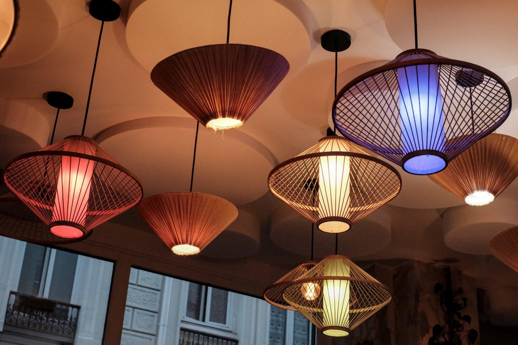 Photo horizontale. Contre plongée sur un plafond décoré de lumière aux applique colorée (rouge, bleu, jaune) en forme de 2 parapluies sans voile posé l’un sur l’autre sur l’ouverture. D’autres lampe ont des abats jour plus classique, mais à l’envers, puisqu’au plafond