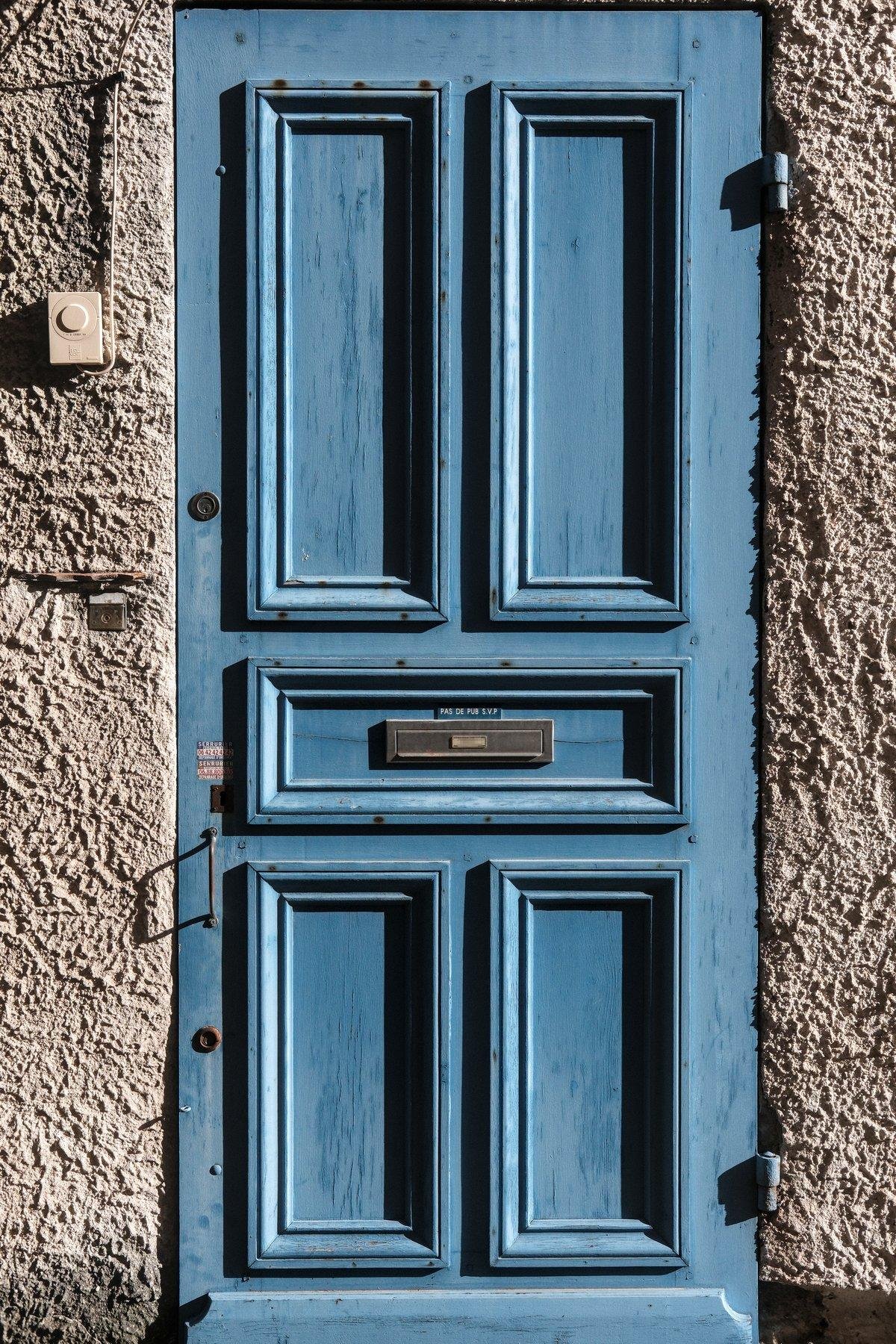 Photo verticale. Une porte bleu dans un mur en crépi. Le soleil éclaire le tout depuis la droite en lumière rasante, faisant ressortir tous les reliefs du mur et les ornements de la porte
