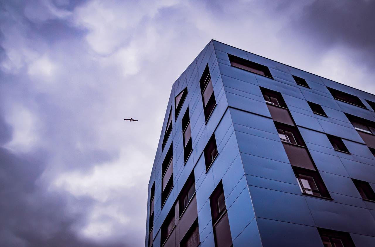 The image shows the corner of a modern, angular building with reflective glass panels. It appears to be a high-rise structure. In the background, there is a cloudy sky, and a bird is silhouetted against it, flying near the top left of the image. The overall mood is somewhat moody and dramatic due to the cloud cover and lighting.
