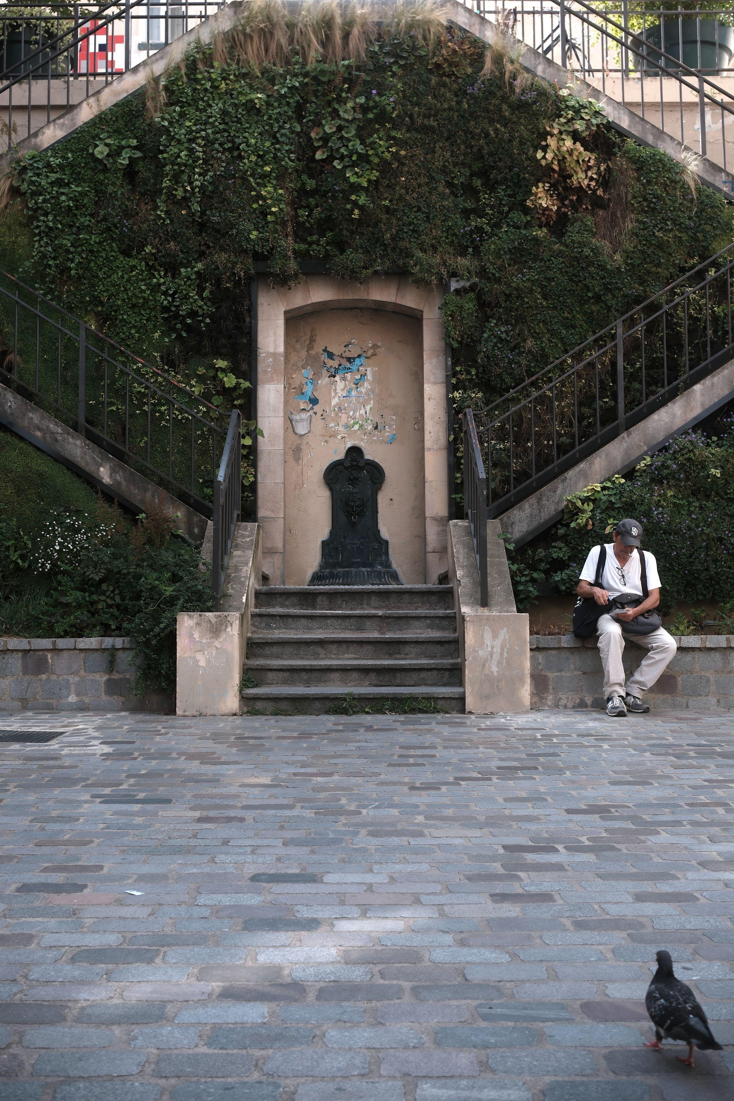 Stone stairs with a metal fountain, surrounded by greenery, plants and stairs on both sides; a man sitting on the right side and a pigeon walking on a cobblestone ground.
