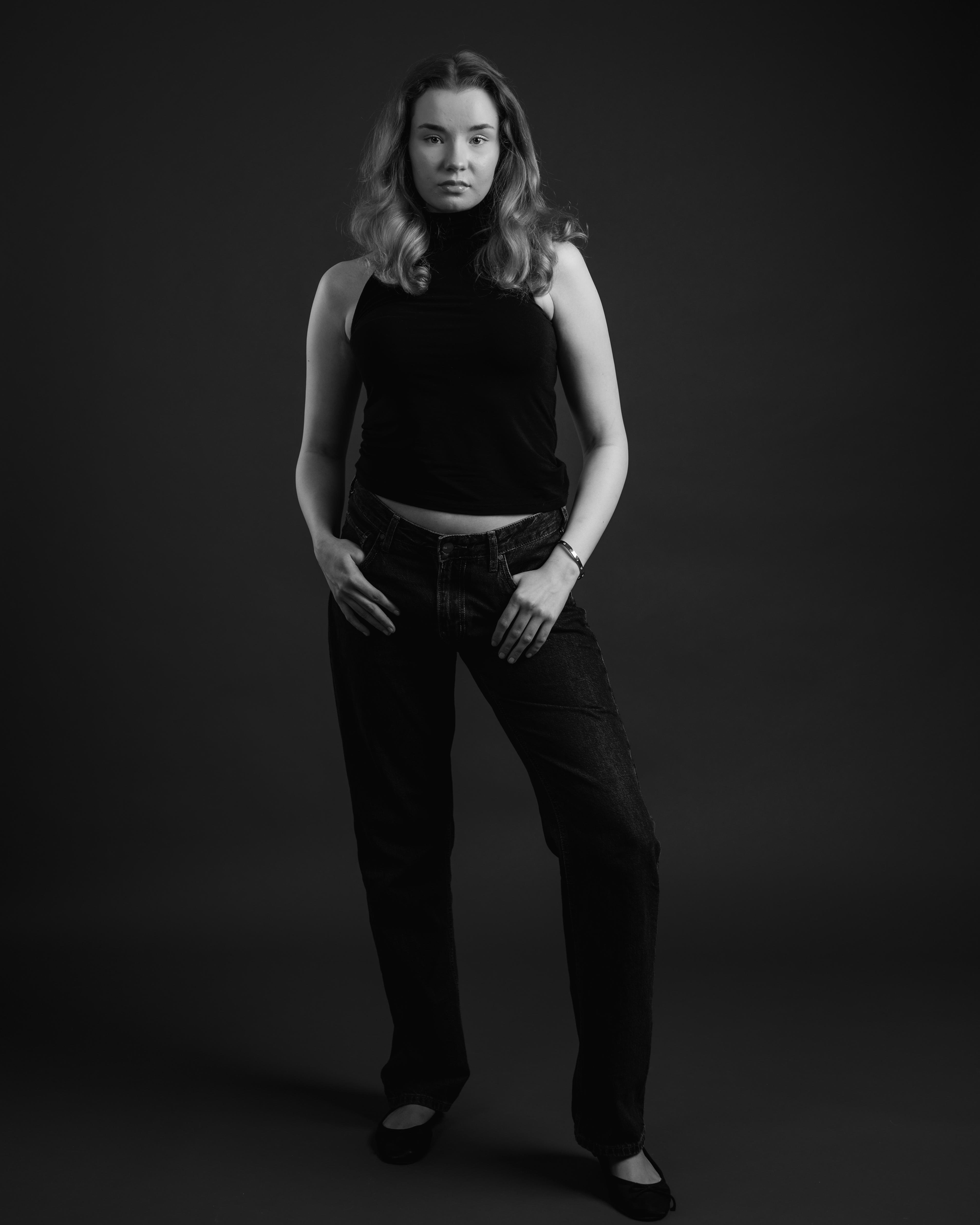 The image is a black and white photograph of a person standing in a studio setting. They are posing confidently, wearing a sleeveless top and jeans. The overall tone is classic and professional. Muotikuvaus. Mallikuvaus Valokuvaaja Kuvaajankulma