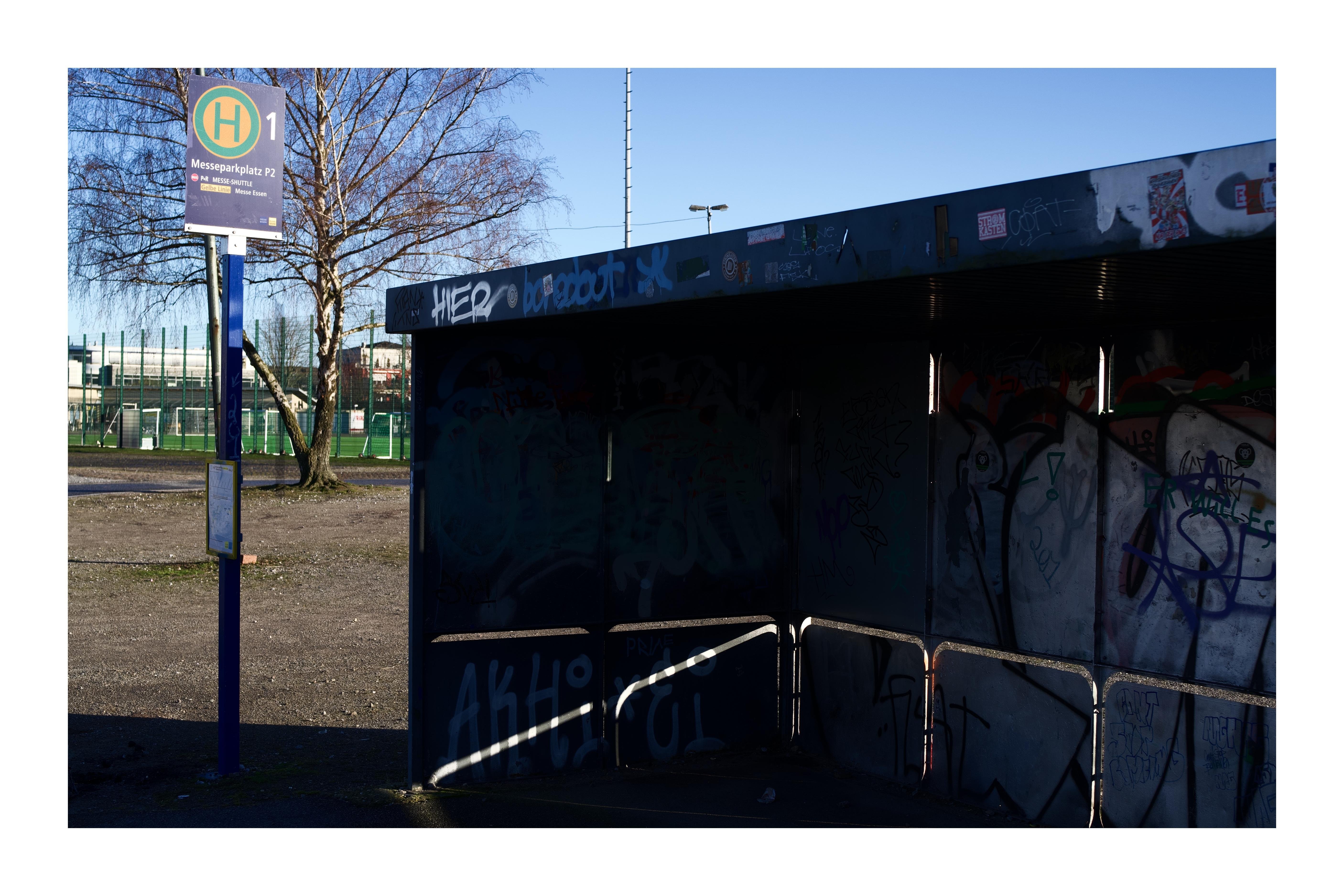 Bushaltestelle „Messeparkplatz P2“ mit graffitiverziertem, teils im Schatten liegendem Wartehäuschen und diagonaler Licht- und Schattenlinie auf der Trennwand der Haltestelle.