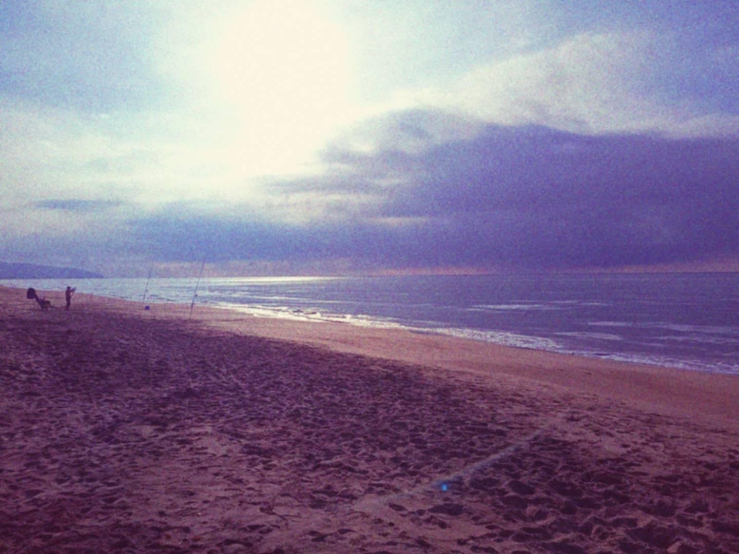 Pescatore su una spiaggia con la sua sedia a sdraio e le canne da pesca piantate nella sabbia, il cielo è nuvoloso.
