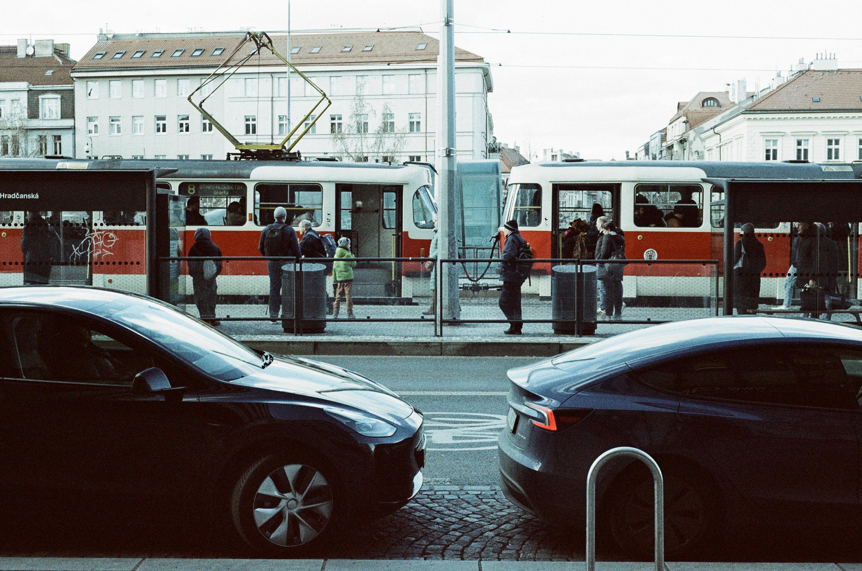 The image shows a street scene with two parked cars in the foreground and a tram in the background. People are waiting at the tram stop. There are buildings visible behind the tram. The scene appears to be taken in an urban area.
