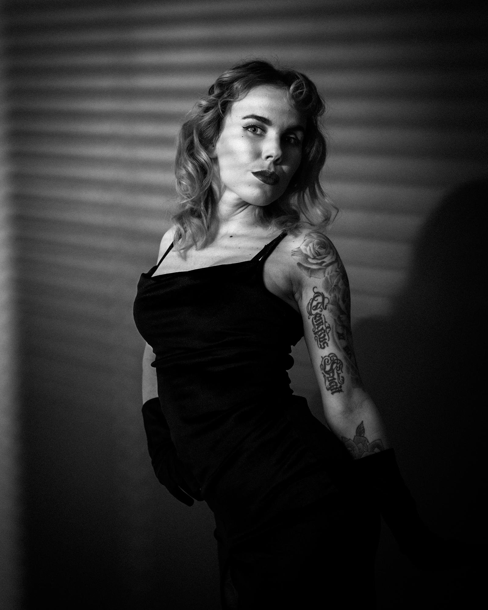 The image is a black-and-white photograph of a person posing with a confident expression. They have shoulder-length curly hair and are wearing a sleeveless outfit. There are tattoos visible on their arm, and they are casting a shadow on the background. The lighting creates stripes on the person and the background, adding to the dramatic effect.