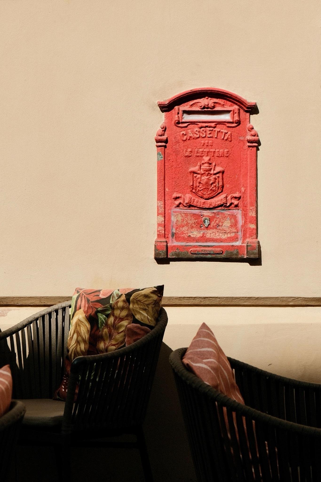 A historic cast iron post box with red painting, set into a beige-yellow wall. The red paint is falling off at exposed spots. The iron front has Italian writing embossed, saying: „Cassetta per le lettere“.
If you look closely, the lid is sealed with transparent silicone, since it is out of use.
In the foreground there are two chairs with colourful cushions, belonging to the Café‘s terrace next door.