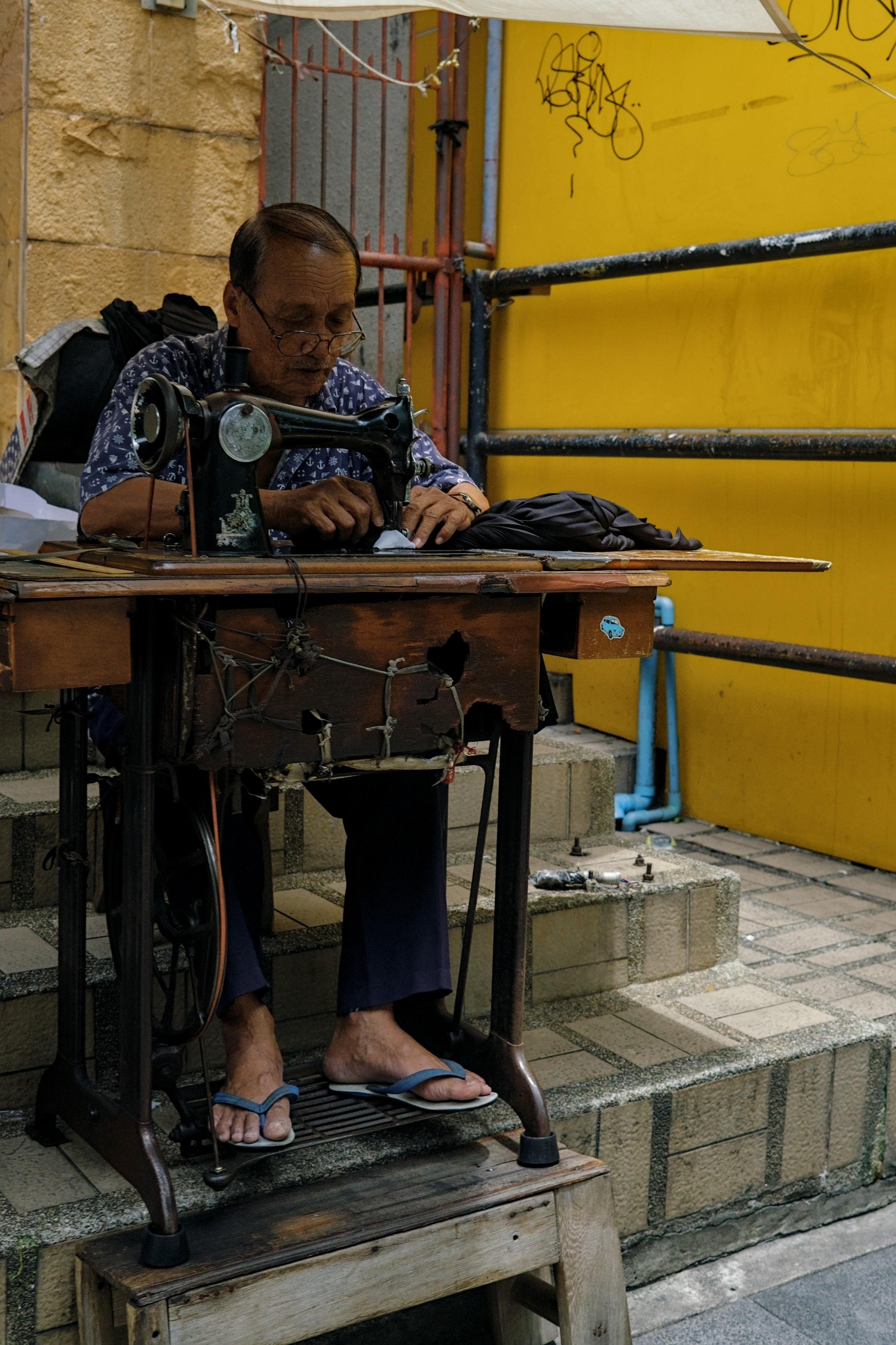 An older man sewing on a manual sewing machine, held together with elastic band, in the middle of the street.