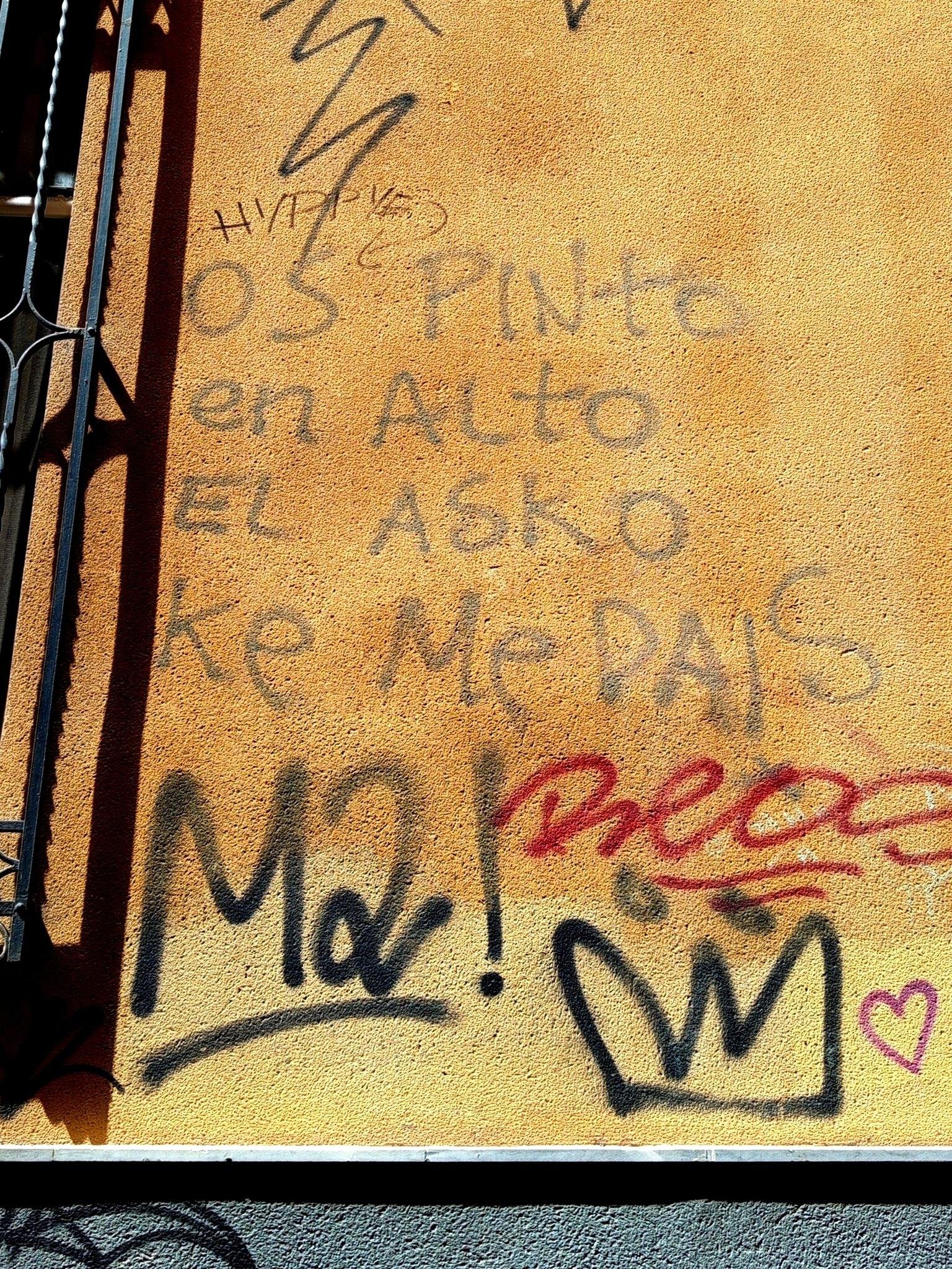 Una pared amarilla de un muro exterior de un edificio con diversos graffitis hechos con aerosoles. En color gris se lee "os pinto en alto el asko ke me dais"