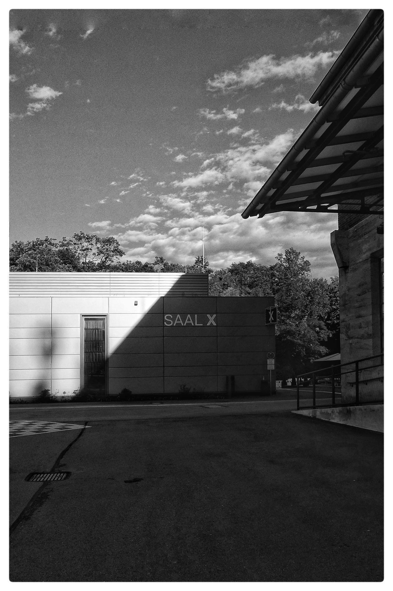 Black and white photo of a modern building with "SAAL X" written on its facade. A large shadow covers part of the building and the pavement in front. The sky is partly cloudy, and trees are visible in the background. Foreground includes part of a sloped roof structure on the right side.