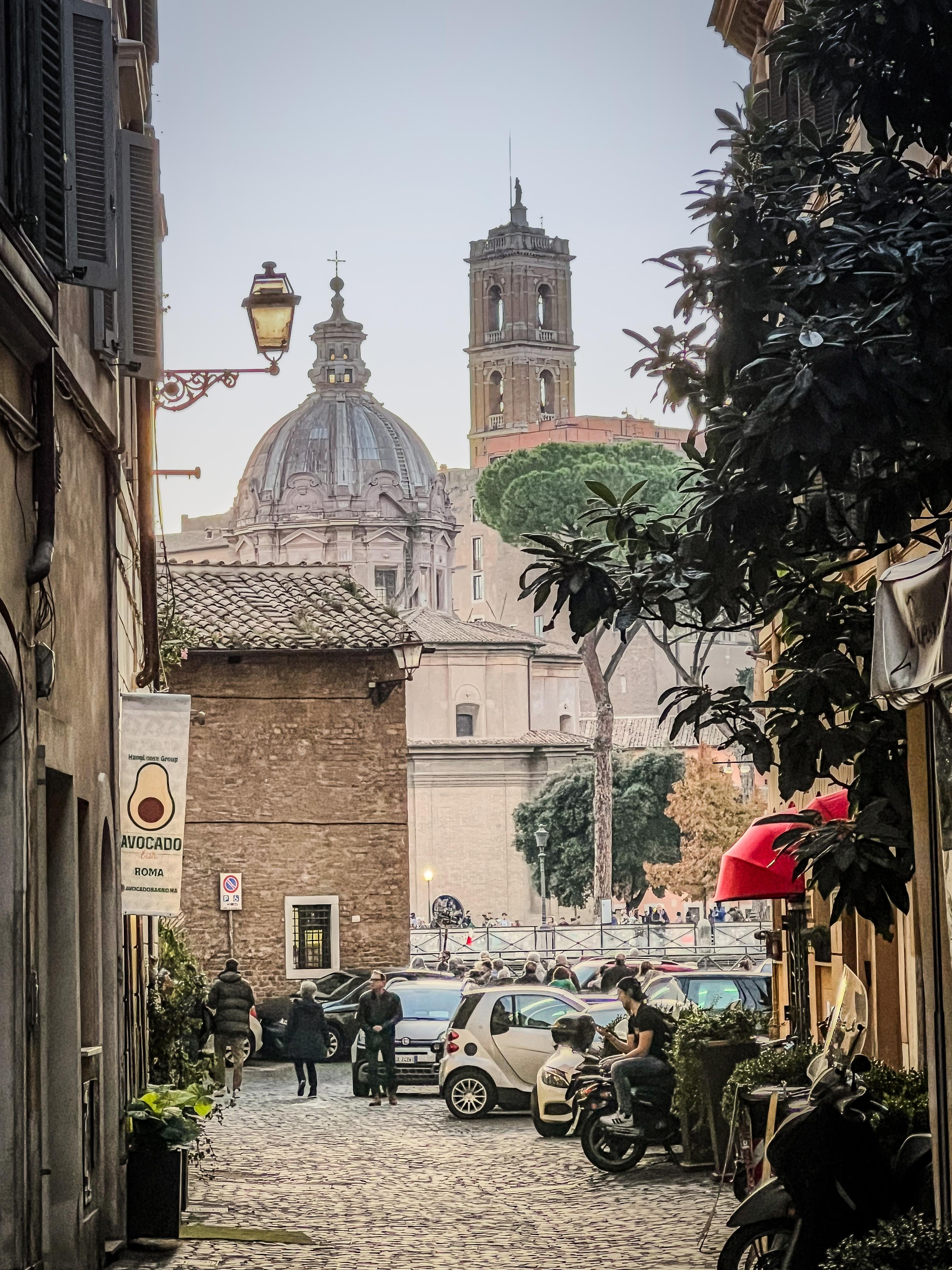 Scena di strada in una città storica europea con pavimento in ciottoli e una vista di punti di riferimento architettonici, tra cui una cupola e un campanile, tipici dell’architettura italiana. C’è anche attività di strada con persone che camminano e auto parcheggiate lungo la strada. Sono visibili insegne e vegetazione, che aggiungono all’atmosfera urbana.