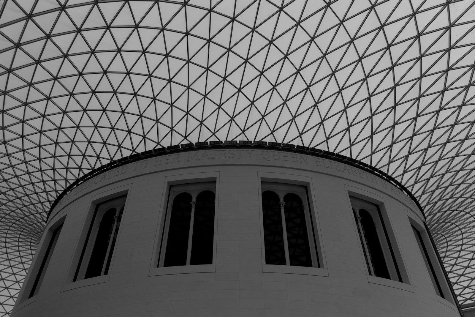 Photo format portrait en noir et blanc, en contre plongée montrant le plafond du hall circulaire du British Museum, et son maillage iconique, avec le bâtiment cylindrique au centre et sa dédicace à la Reine Elizabeth II