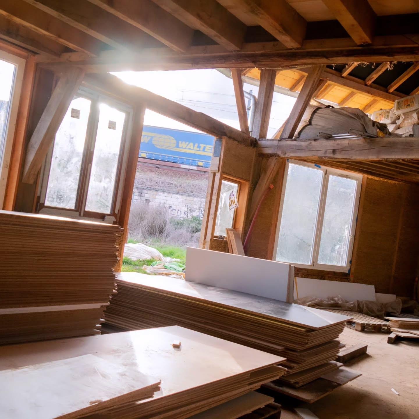 The image shows the interior of a building under construction or renovation. There are stacks of wooden boards or panels on the floor. The structure has large windows, and natural light is coming in. The walls and ceiling appear to be made of wood, indicating an unfinished or ongoing construction project. Outside, there is greenery and part of a freight train with a blue and yellow design visible through the window.