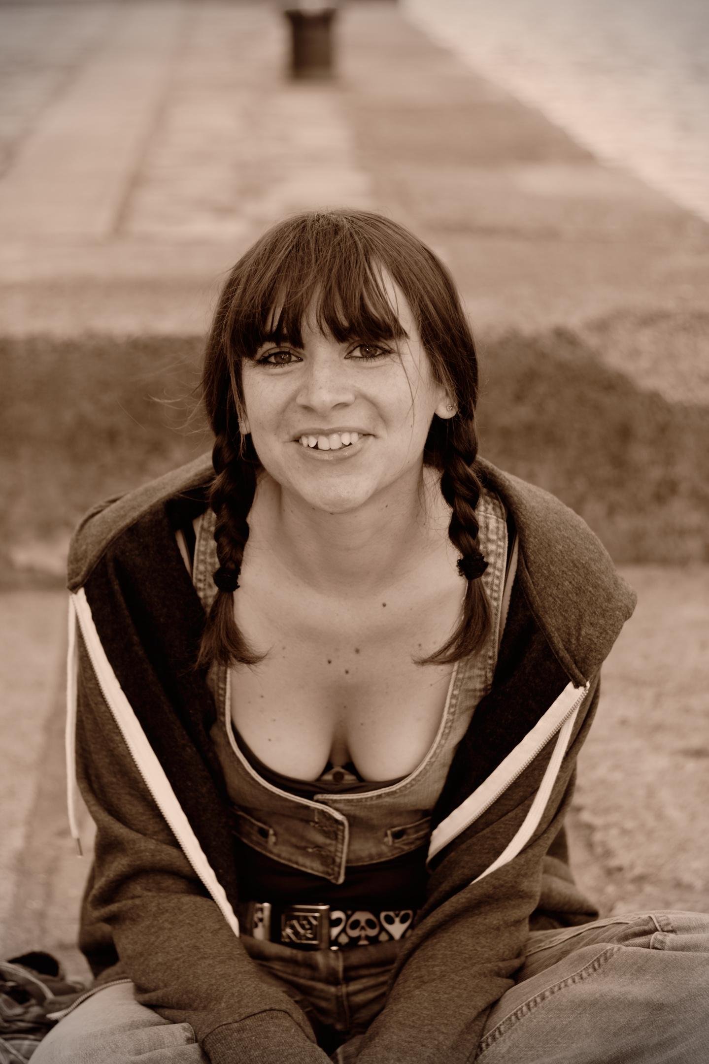 Photo verticale en sépia. Une jeune femme assise presque en tailleur (jambes descendent une marche) Elle est en débardeur, avec par dessus un gilet sans manche puis un hoodie à zip ouvert. Elle a deux nattes de part et d’autre du visage. Elle regarde la caméra en souriant.