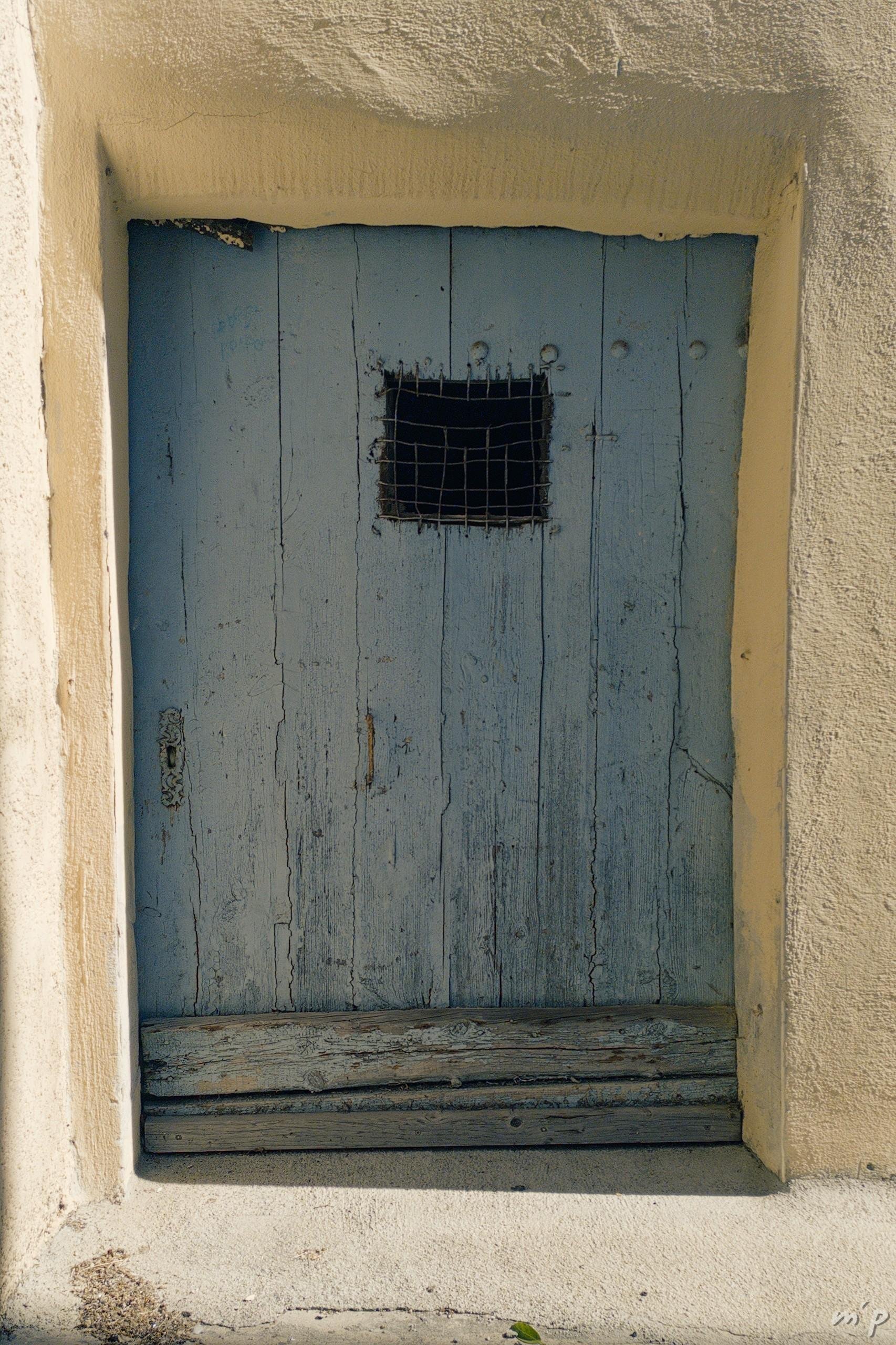 🇬🇧 Vertical color photo of a small low door of faded and worn blue wood with an opening in the upper part carrying an irregular grille. The frame is covered with an irregular light sand-colored coating. The sun casts some shadows.

🇫🇷 Photo couleur verticale d'une petite porte basse de bois bleu délavé et usé avec une ouverture dans la partie supérieure portant une grille irrégulière. L'encadrement est recouvert d'un enduit irrégulier couleur sable clair. Le soleil projette quelques ombres.