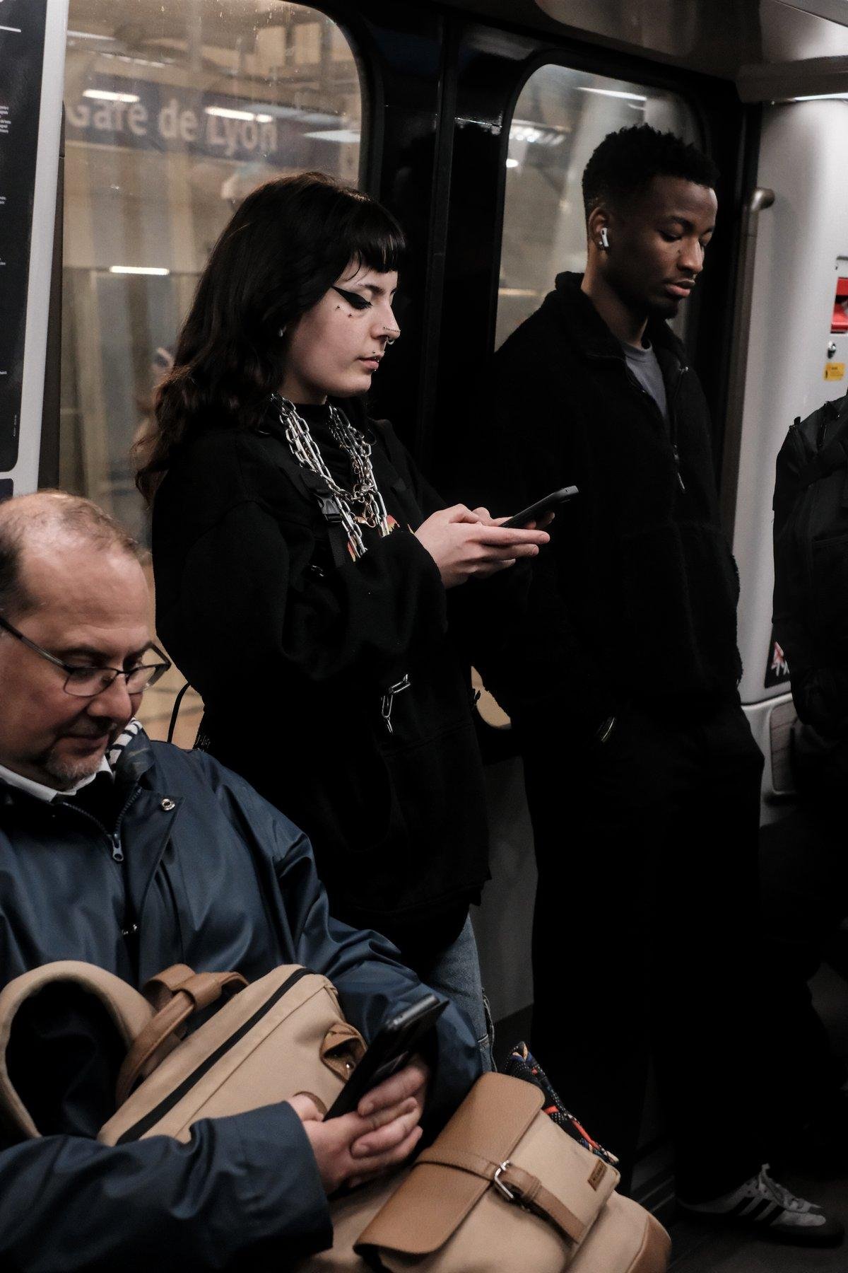 Photo verticale. Une jeune femme brune au cheveux longs, adossée à la double porte du wagon du métro, entrain do consulter son téléphone. Elle porte un haut noir à manches longues, de grosses chaînes en acier autour du cou, un maquillage noir marqué sur les yeux.