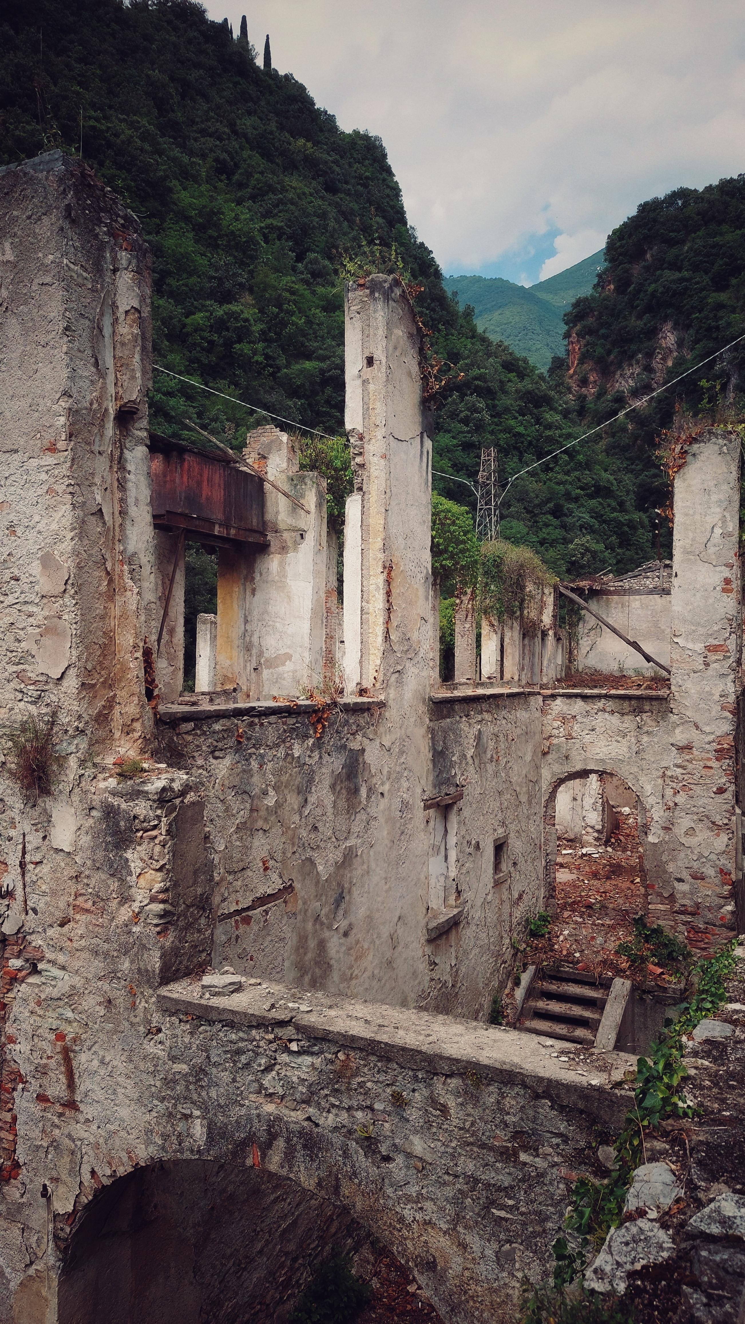 Ruins of an old stone building with partially collapsed walls and arches, surrounded by lush green forested hills. The sky is partially cloudy with patches of blue. Vegetation grows atop the walls and cracks, indicating age and abandonment. A rusted beam and power line cross the scene, adding to the derelict atmosphere.