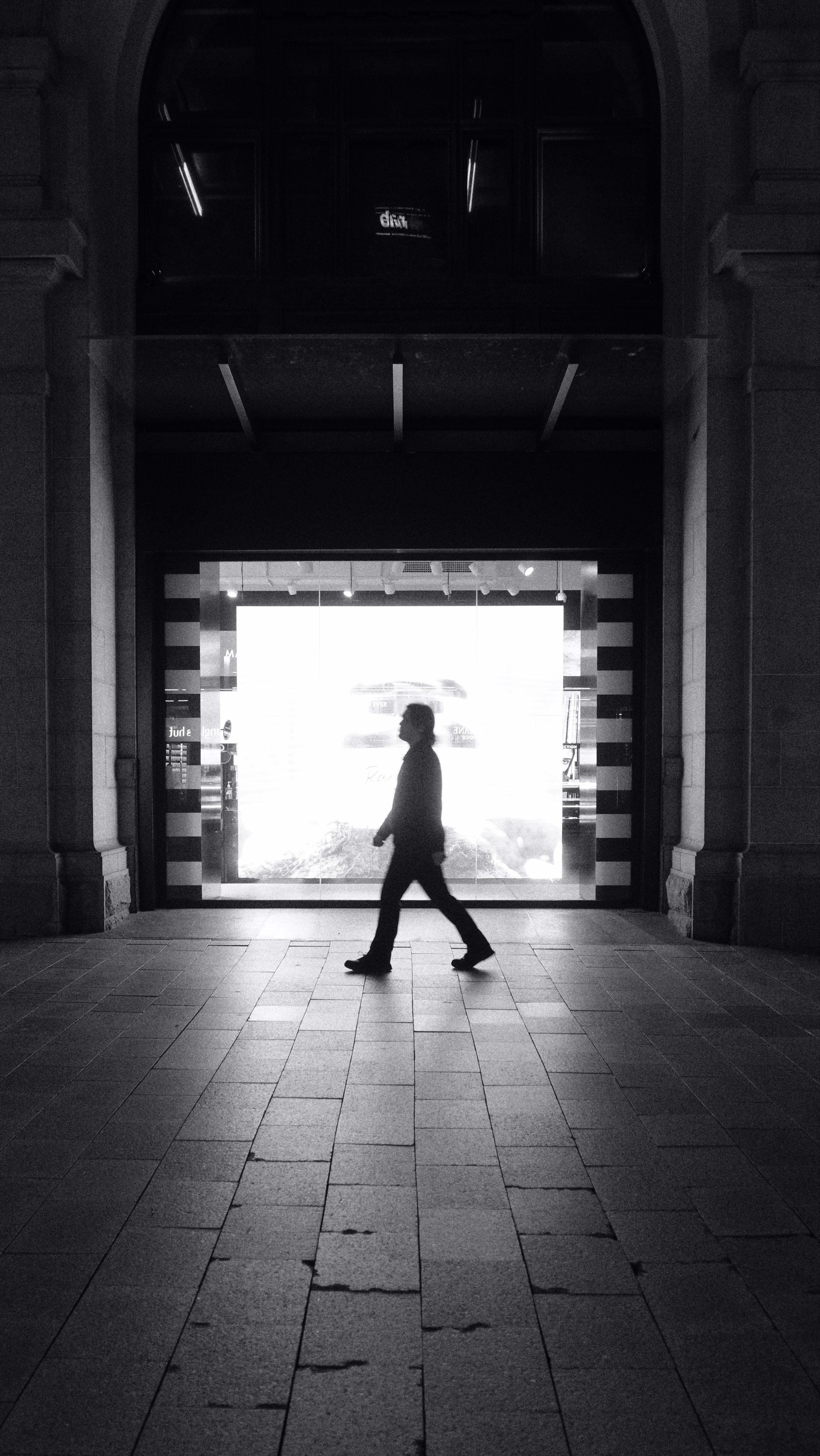 Silhouetted person walking across a tiled footpath in front of a tall stone archway, with a bright illuminated storefront window in the background.