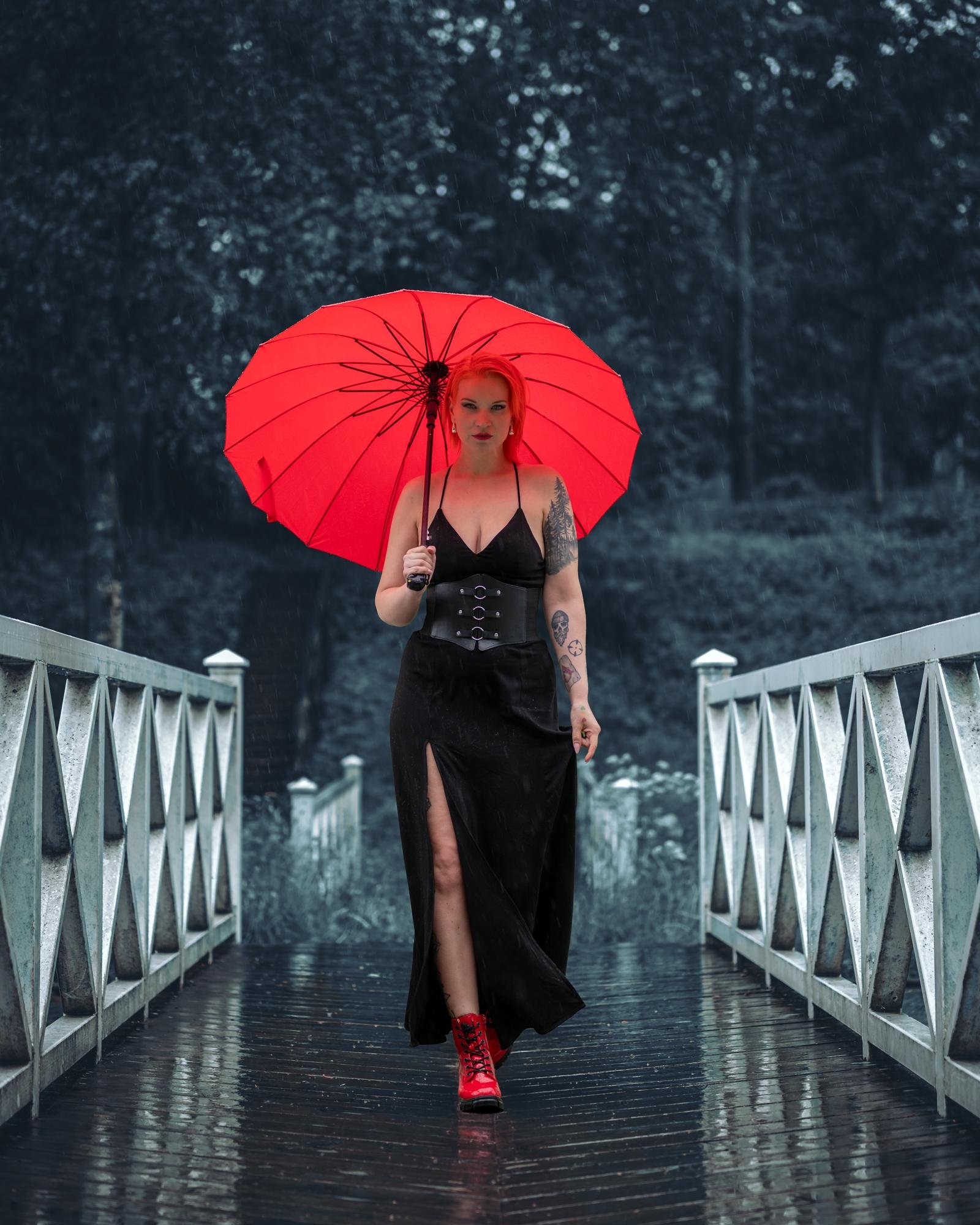 Woman with red hair and tattoos walks down a wet wooden bridge holding a red umbrella. She is wearing a long black dress with a slit and red boots. The background features a blurred view of trees in a rainy, overcast setting.