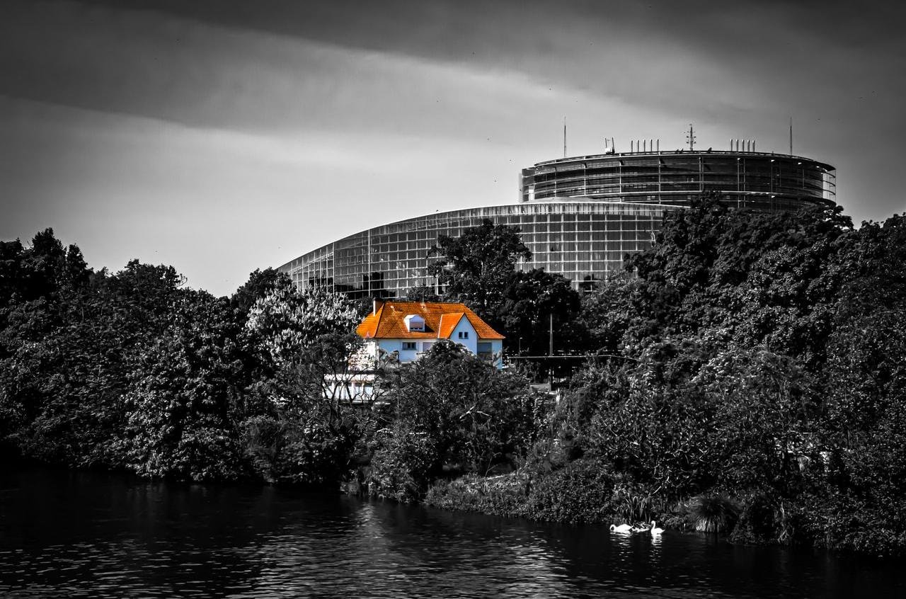 A vibrant orange-roofed house stands out in a black-and-white scene surrounded by dense trees near a body of water. In the background, a large circular, modern building with a glass facade rises above the treetops under a cloudy sky. A pair of white swans is visible on the water in the foreground.
