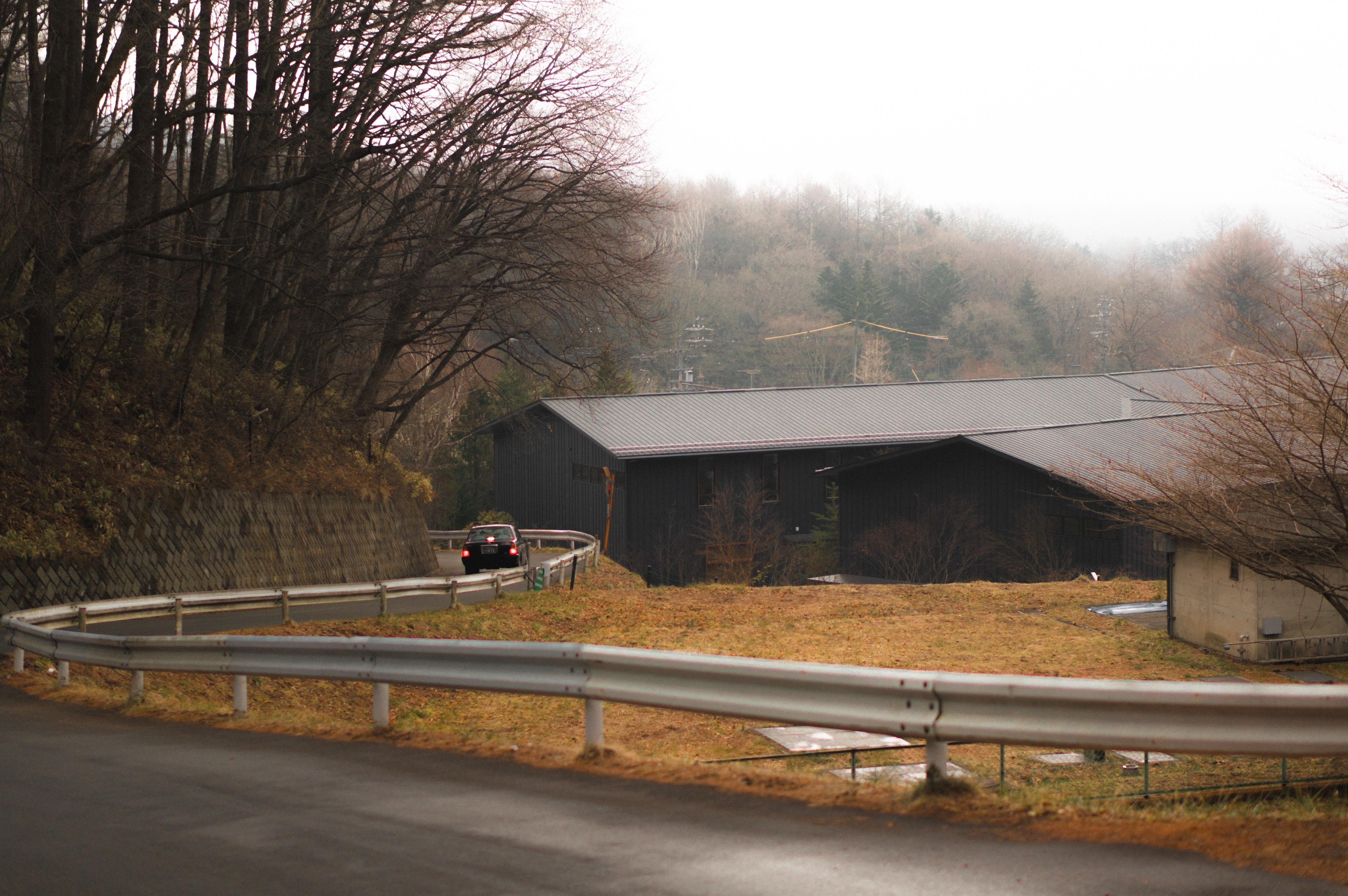 The image features a winding road with a car driving along it, surrounded by trees. There's a field to the right with a large, dark building in the background. The scene has an overcast sky, suggesting a cloudy or foggy day.